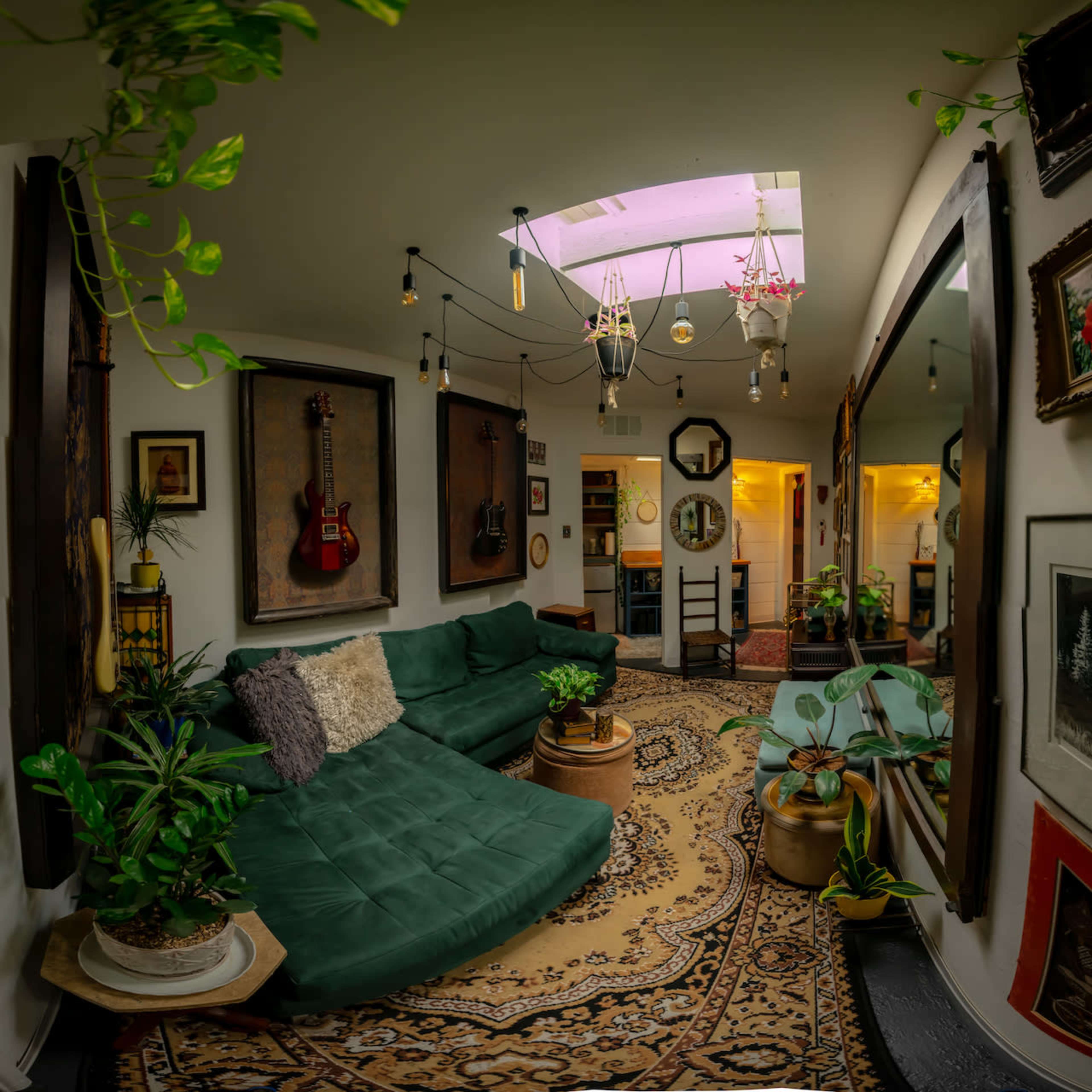 The image shows a cozy living room featuring a green sectional sofa, framed artwork on the walls, plants in various pots, and a skylight illuminating the space.
