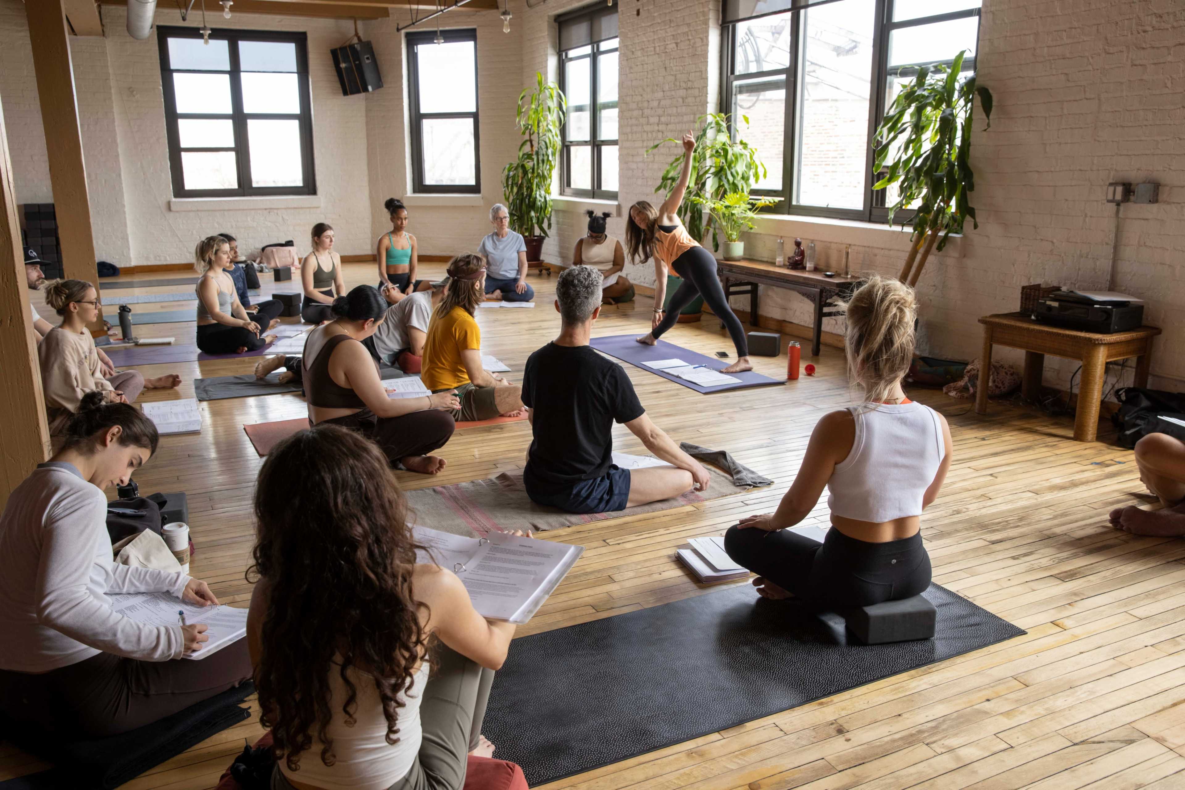 A group of people participates in a yoga class in a spacious studio with large windows and wooden flooring.