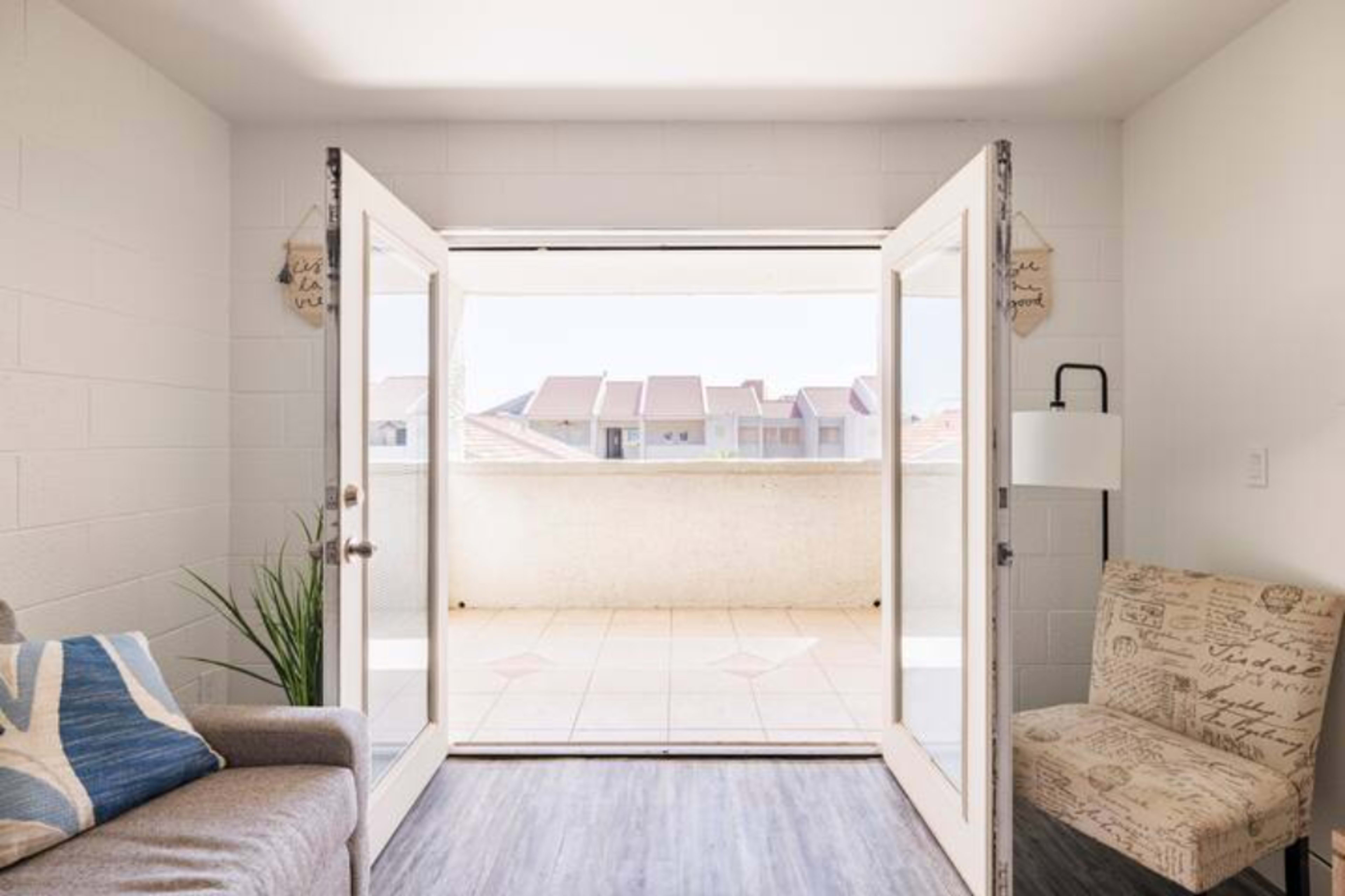 A pair of open French doors leads from a room to a tiled balcony overlooking a multi-story building.