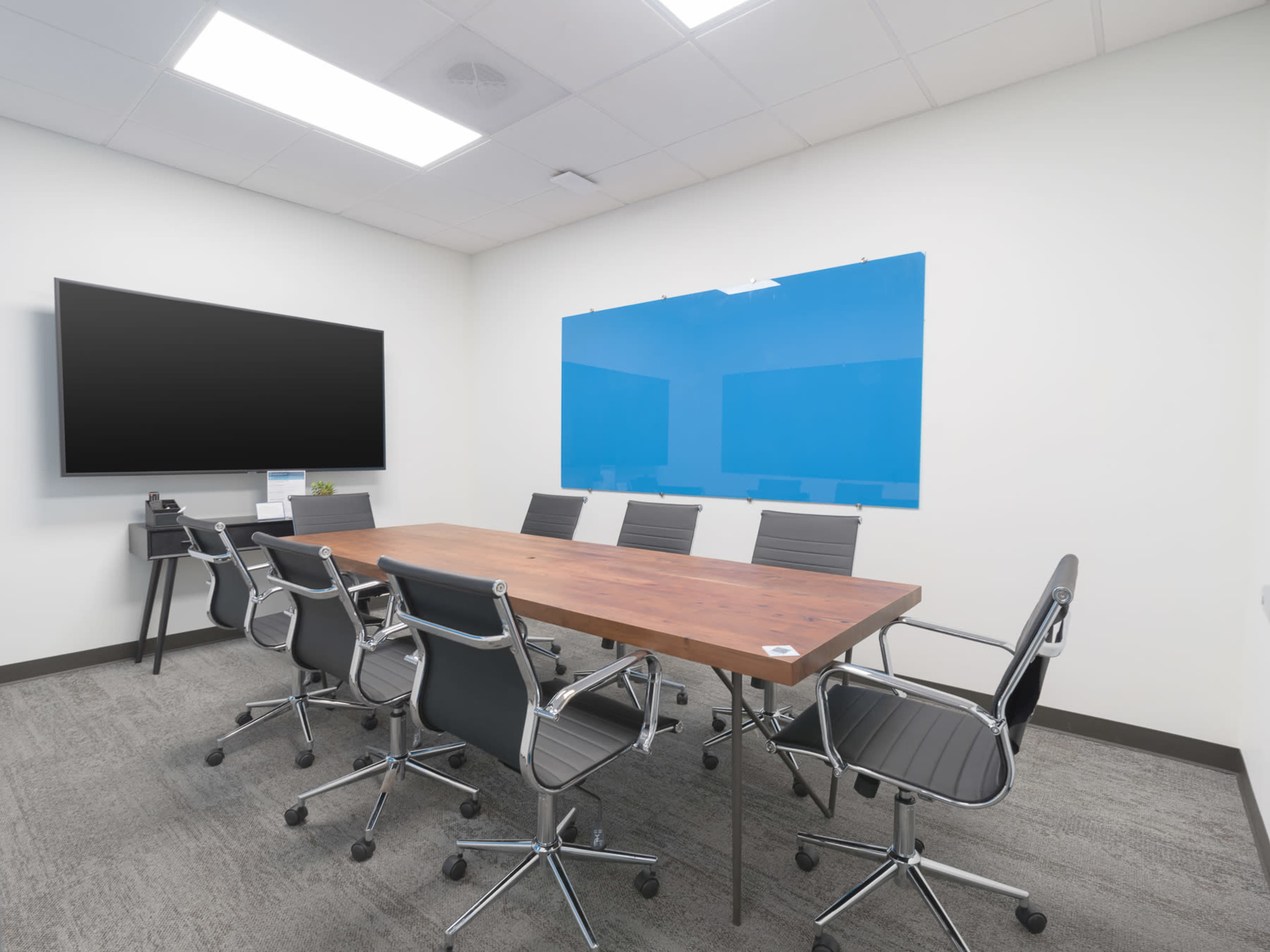 A conference room features a large wooden table surrounded by ergonomic chairs, with a mounted TV and a blue whiteboard on the wall.