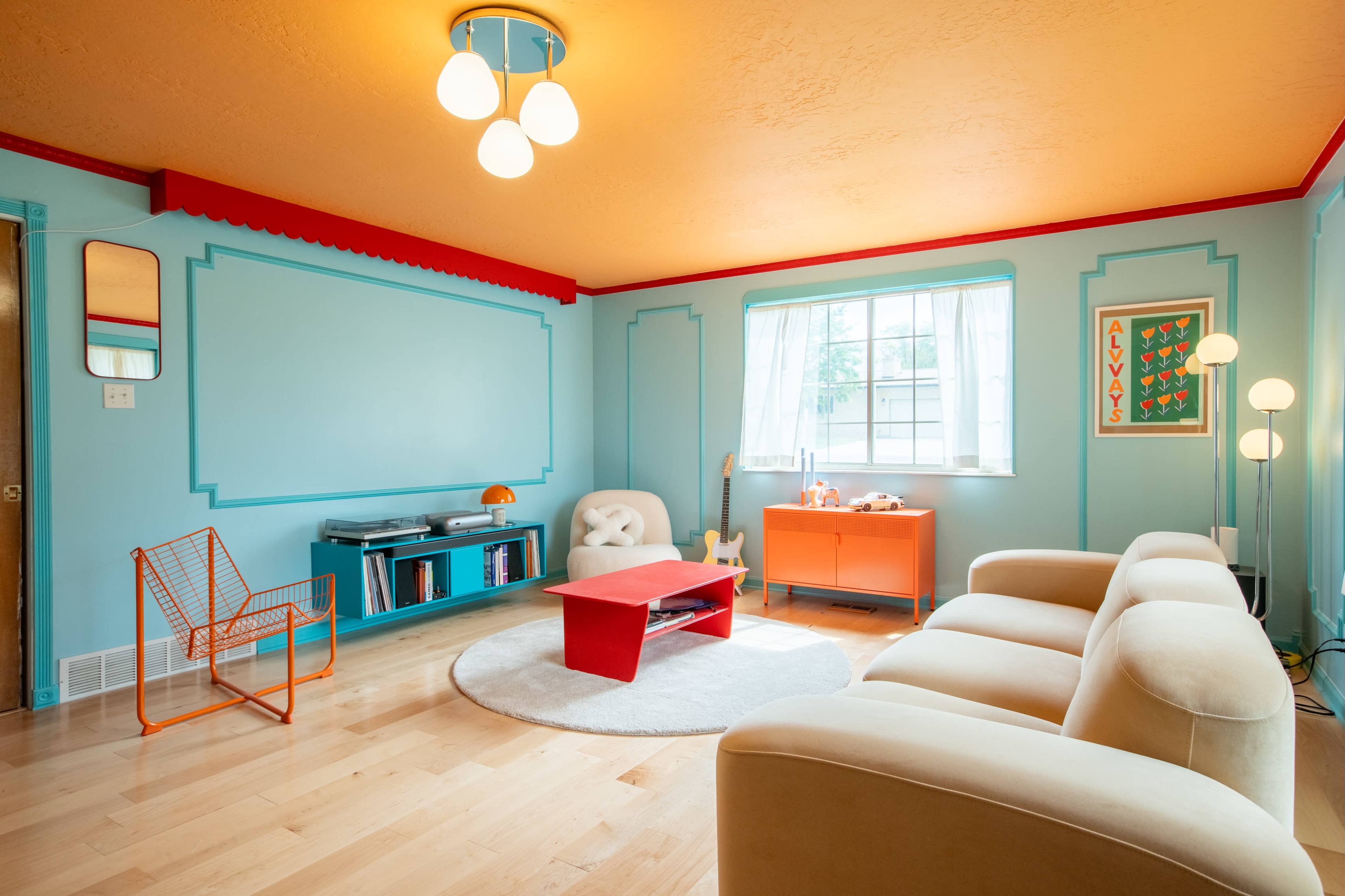 The room features a light blue wall with orange accents, a circular white rug, a red coffee table, and modern furniture.