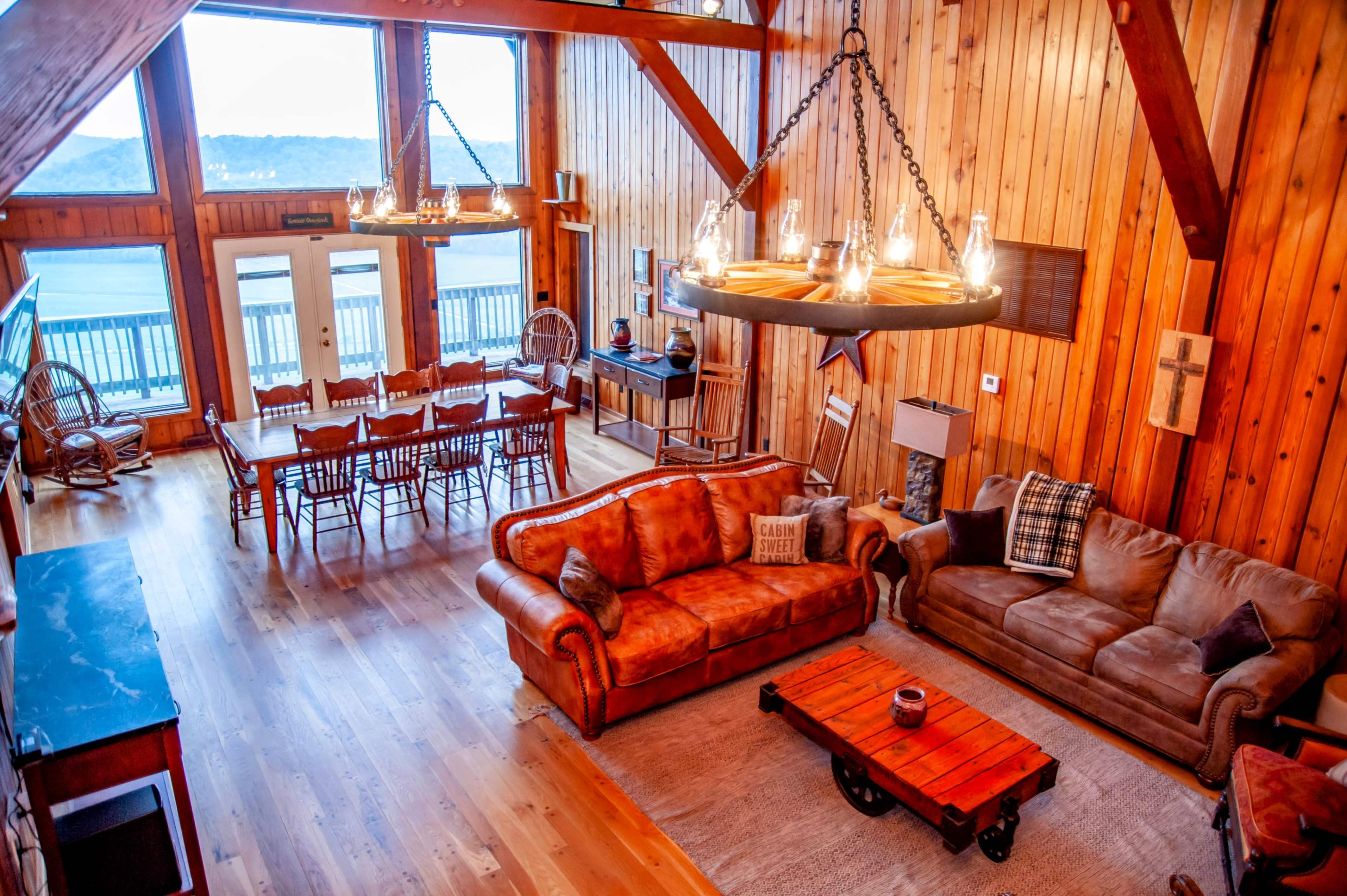 A spacious wooden interior featuring a living area with two sofas, a large dining table, and large windows overlooking a body of water.