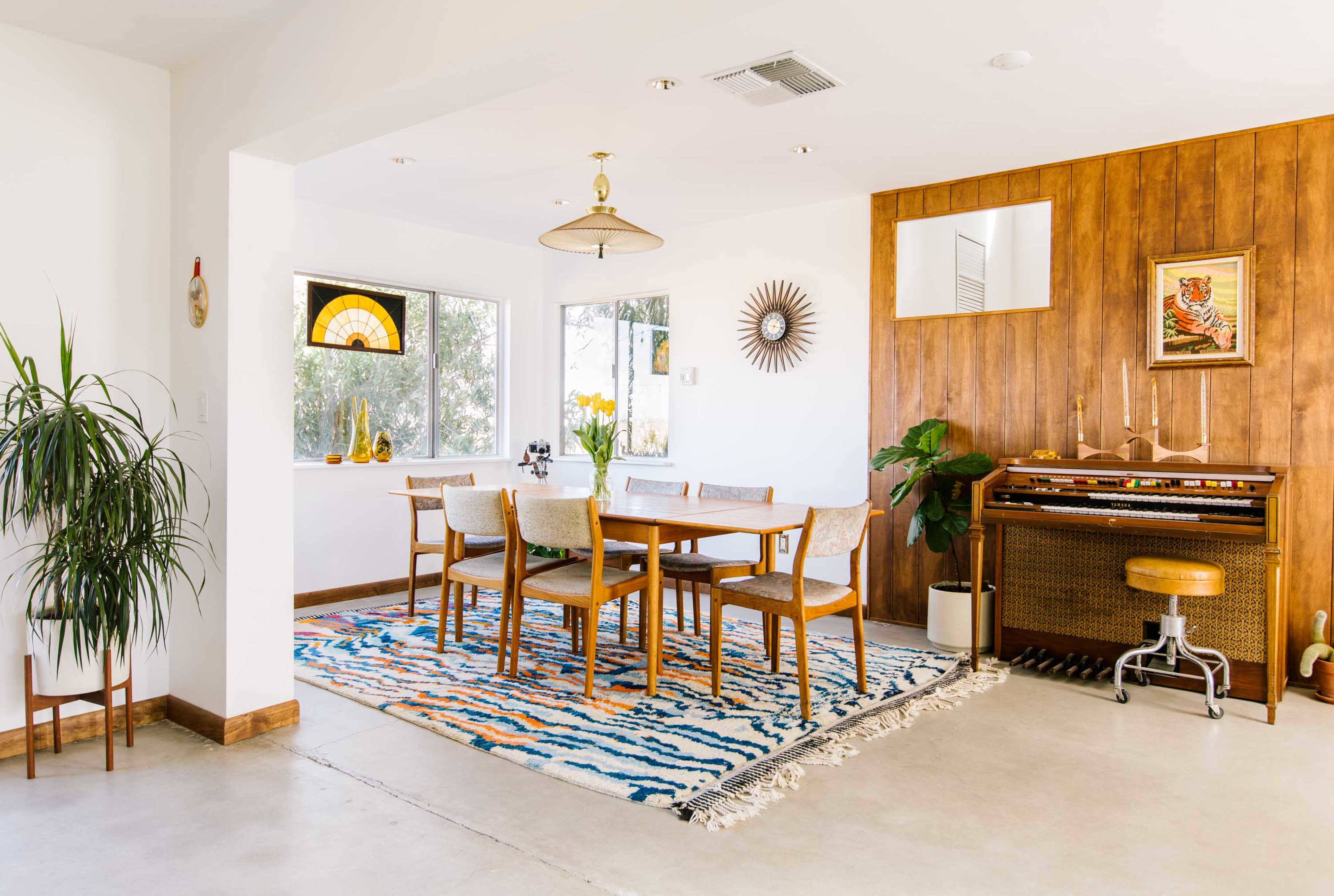 A modern dining area features a wooden table surrounded by six chairs, with a patterned rug beneath and a vintage organ against a wooden wall.