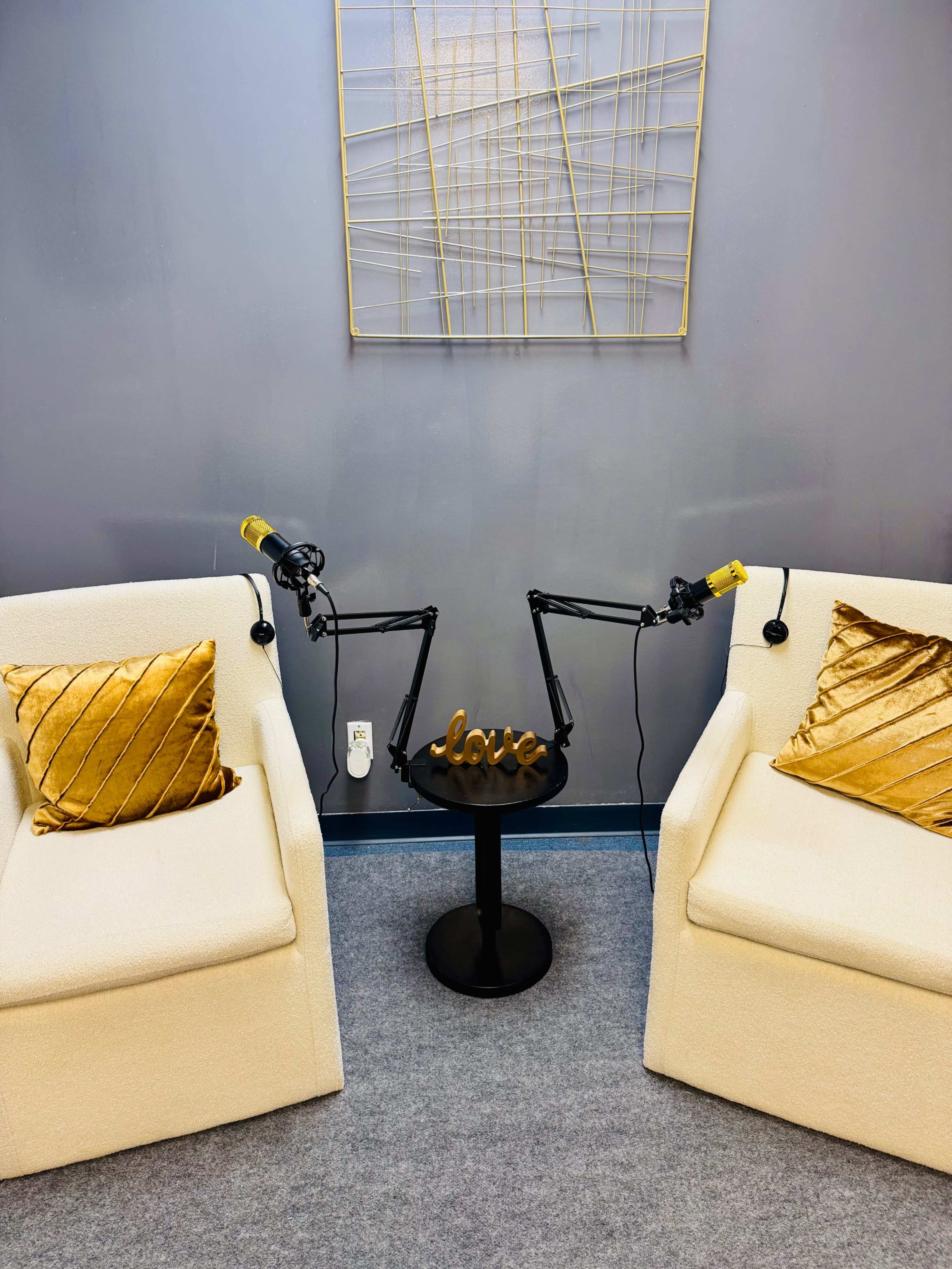 The image shows two cream-colored chairs with golden accent pillows, positioned around a black table featuring a decorative piece that reads "love," and two microphones mounted on adjustable arms.