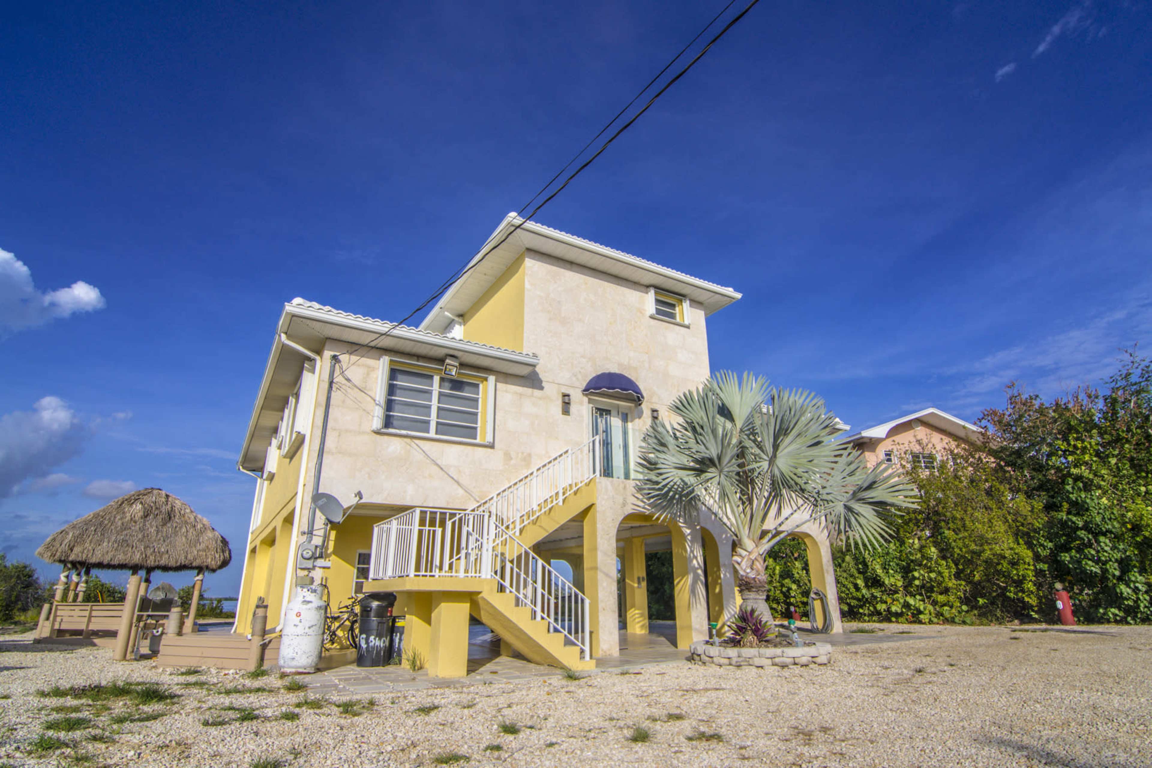 A two-story yellow house with a palm tree in front and a thatched roof structure nearby sits on a gravel driveway under a clear blue sky.