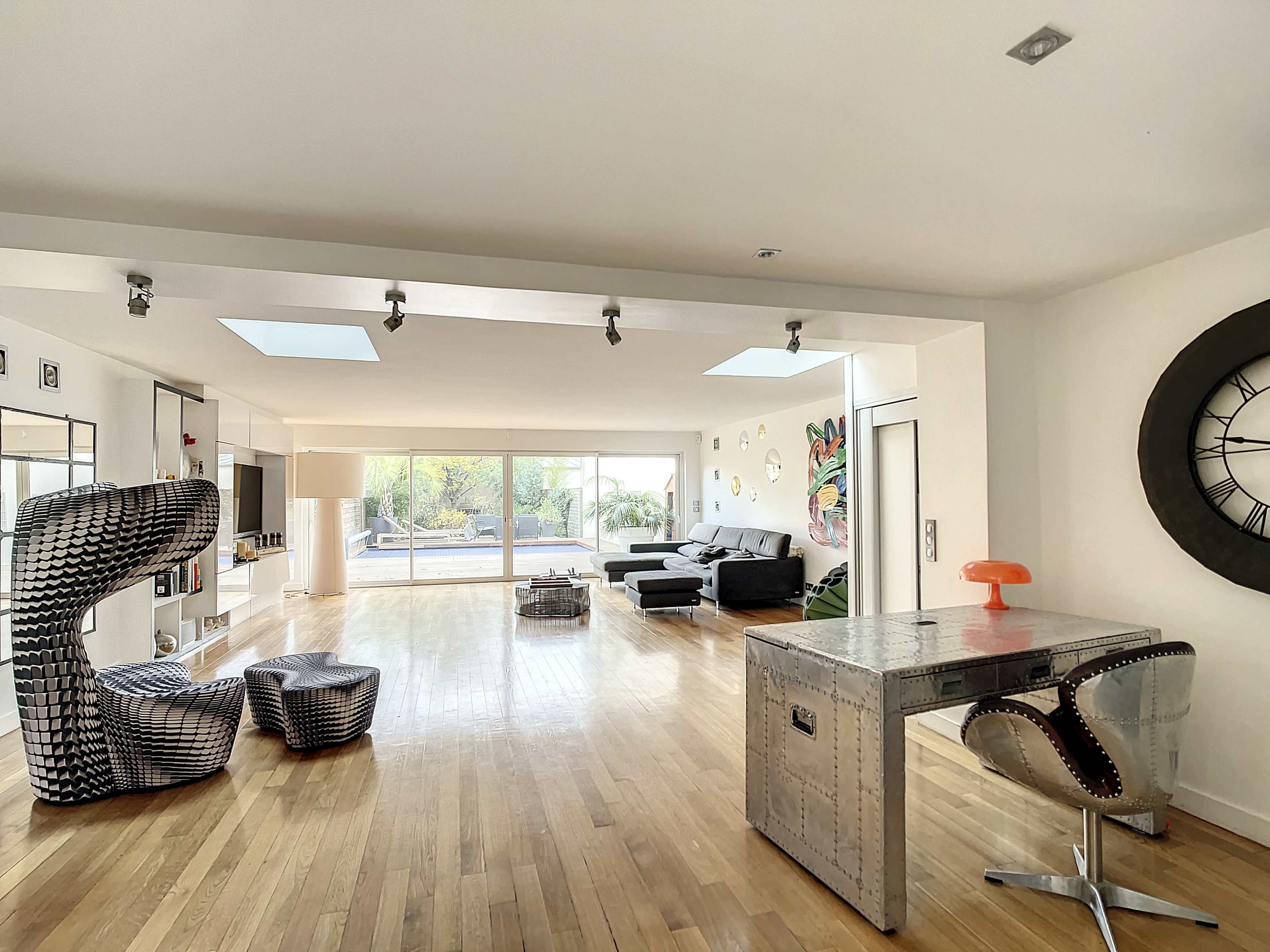 A spacious, modern living area with wooden flooring, a large window allowing natural light, and contemporary furniture pieces, including a unique chair and a metallic table.