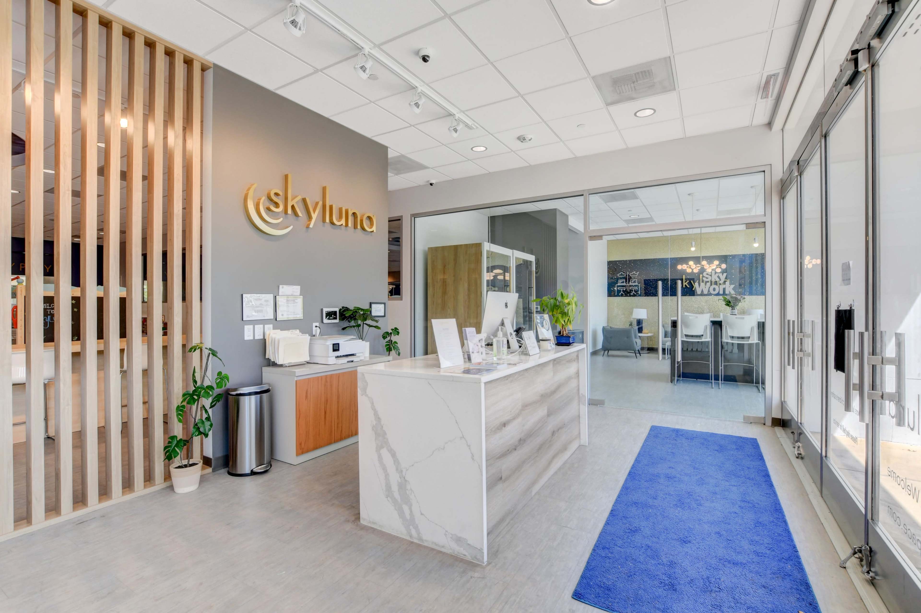 A modern office reception area with a marble front desk, greenery, and a blue welcome mat, featuring a sign that reads "skyluna."