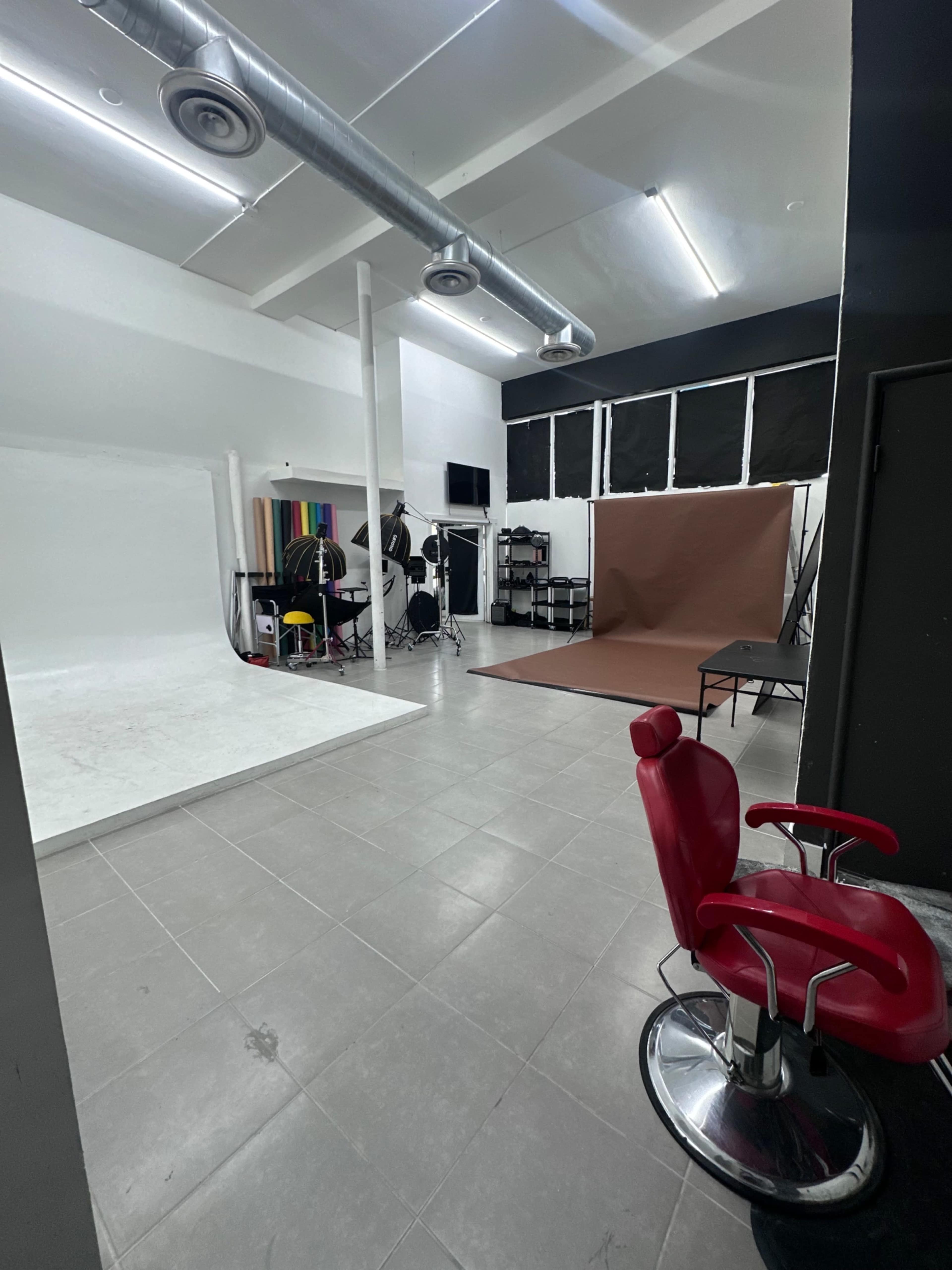 The image shows an empty studio space with a red barber chair, a white backdrop, various equipment, and a brown background on one side.