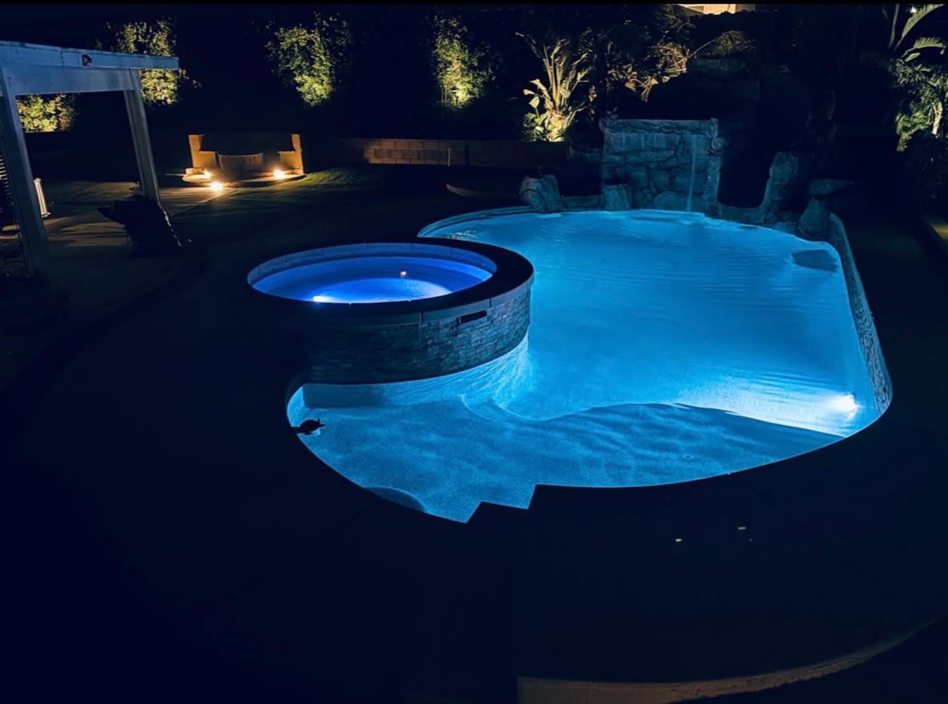 The image shows a swimming pool with a hot tub, illuminated by blue lighting, surrounded by landscaping at night.