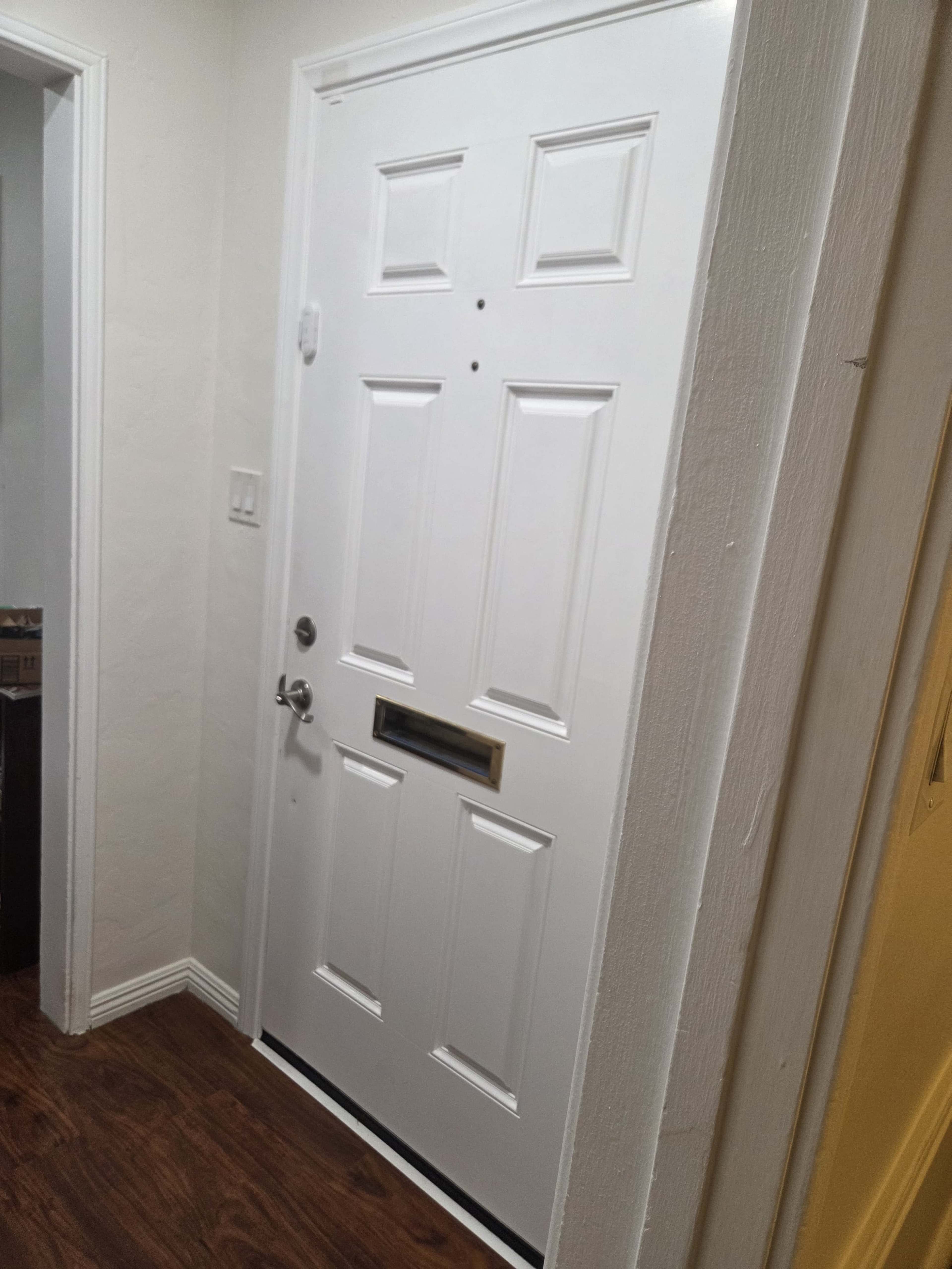 The image shows a white front door located at the end of a narrow hallway, with wooden flooring and a yellow wall visible to the right.