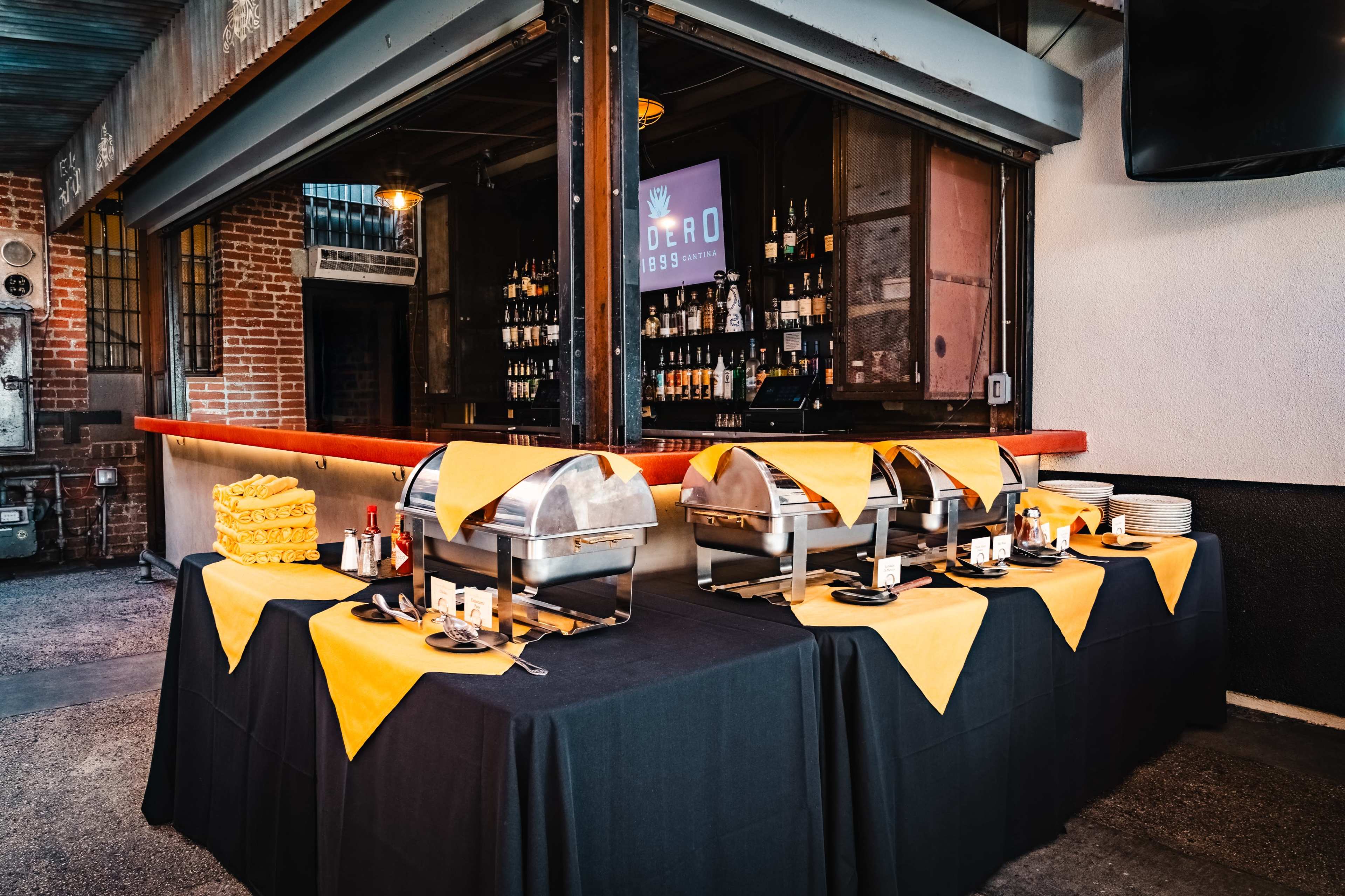 A buffet setup features several chafing dishes on a black tablecloth adorned with yellow table runners, with a bar area visible in the background.