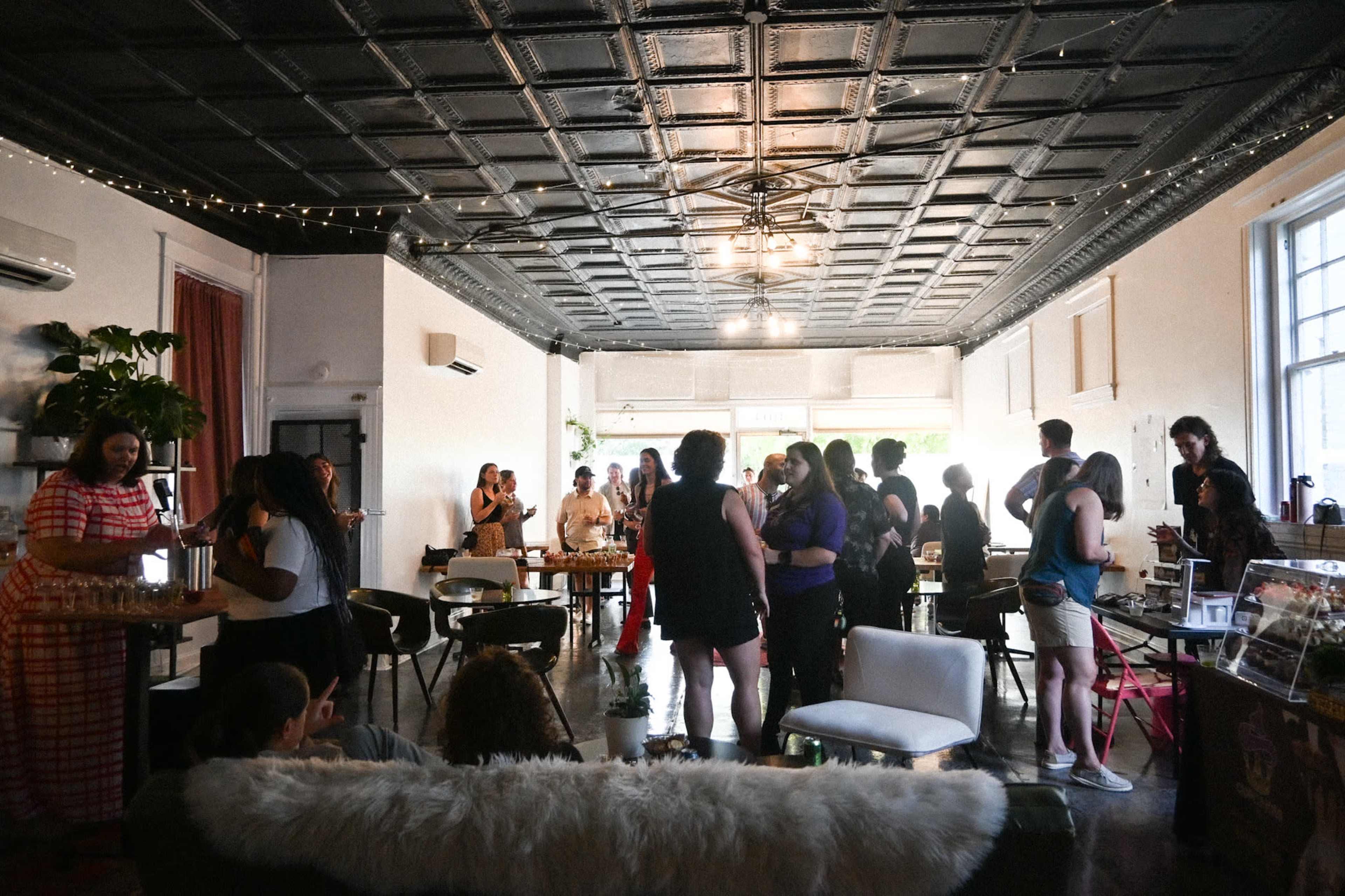 A group of people socializes in a brightly lit room with a patterned ceiling and various tables and chairs set up for seating.