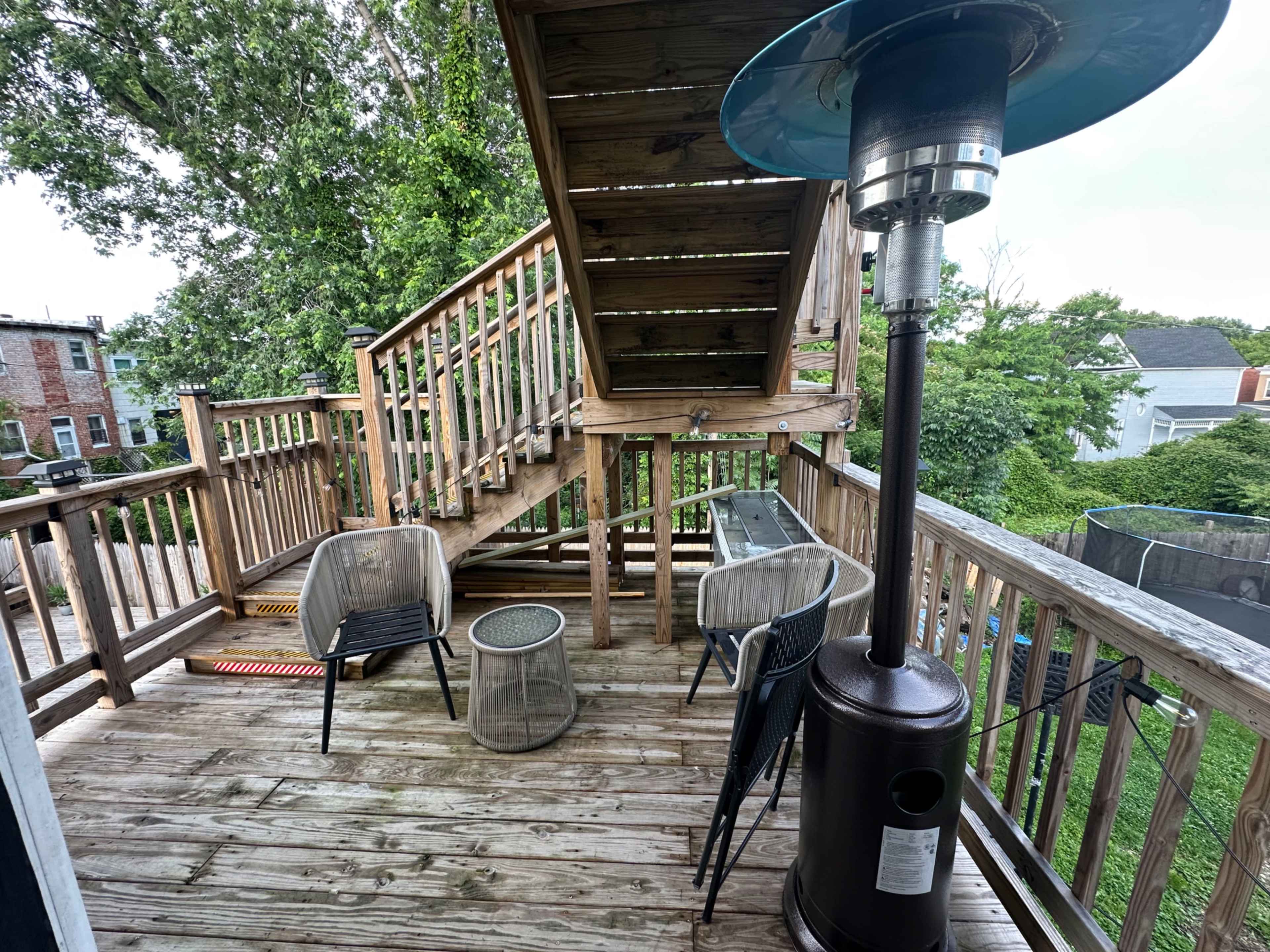 The image shows a wooden deck featuring two chairs, a small table, a patio heater, and a staircase leading to another level.