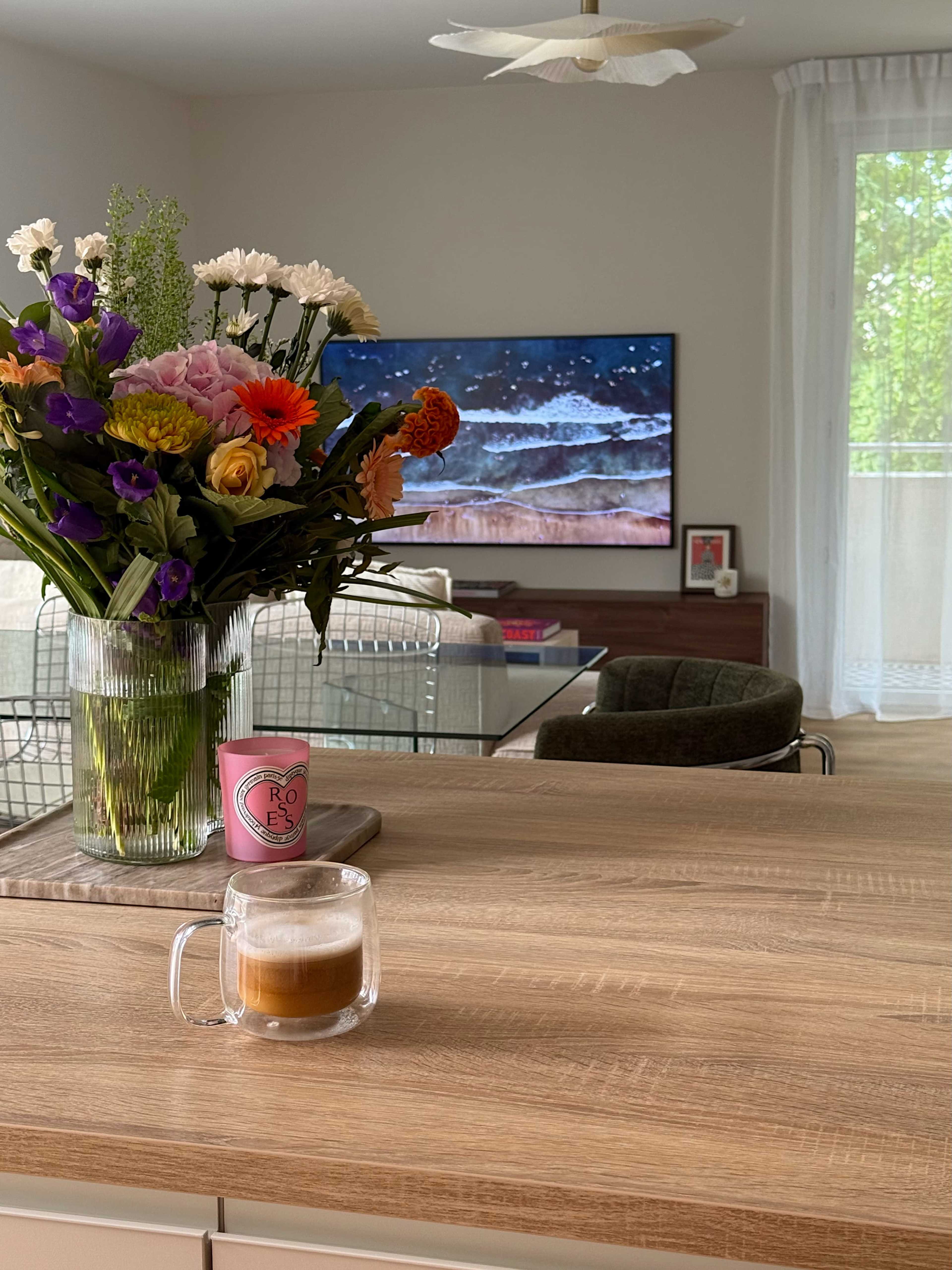 A clear glass mug of coffee sits on a wooden kitchen counter in front of a vase of flowers and a television displaying ocean waves.