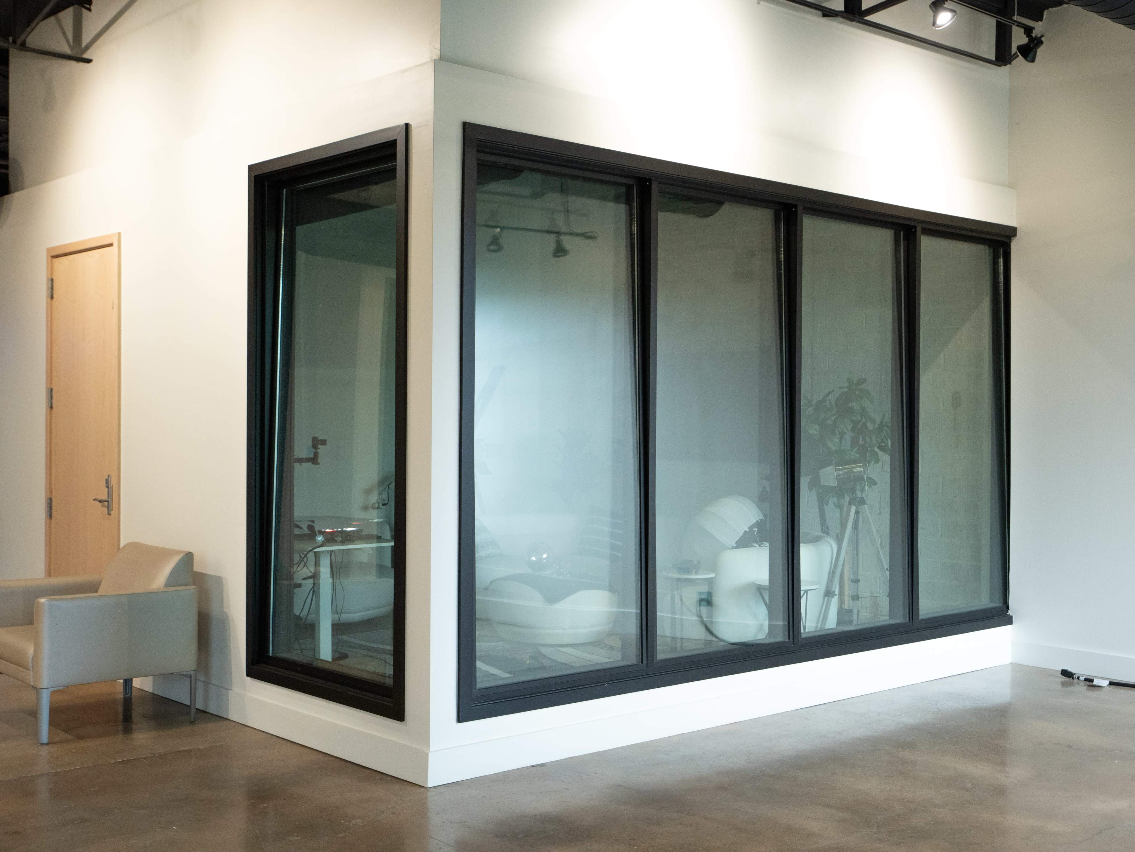 The image shows a modern meeting room enclosed by large glass panels, featuring a chair outside and minimalistic decor inside.