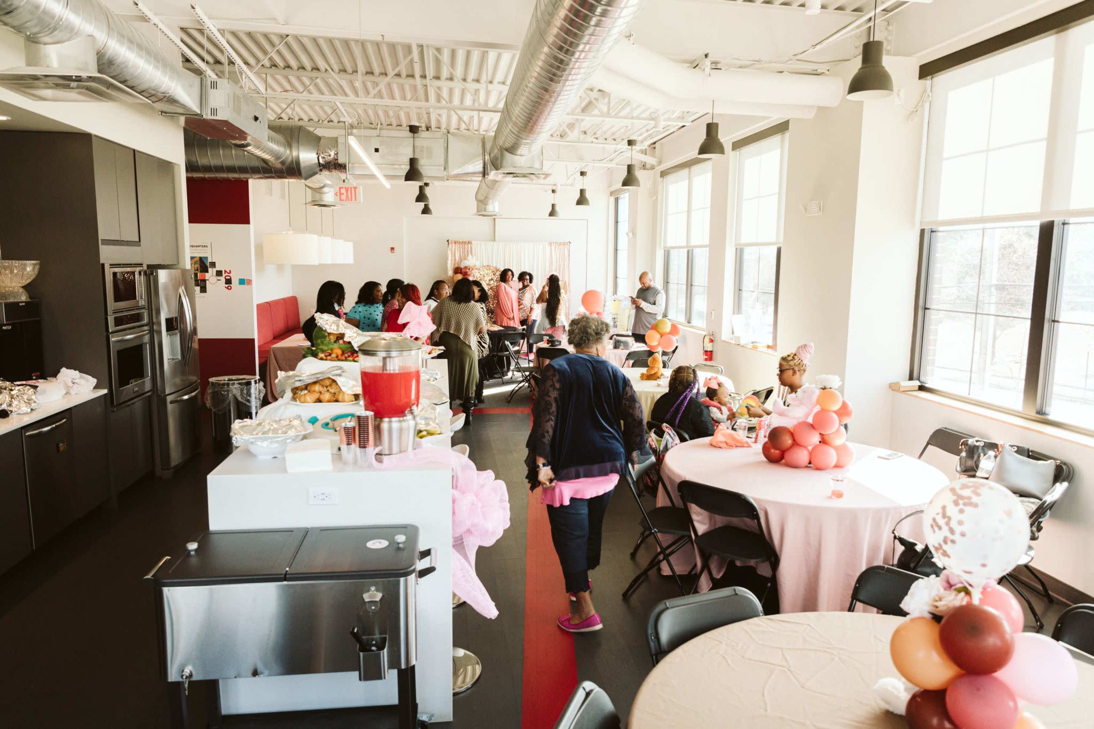 A brightly lit indoor space set up for a gathering, with tables adorned with decorations, food displayed, and guests interacting in the background.