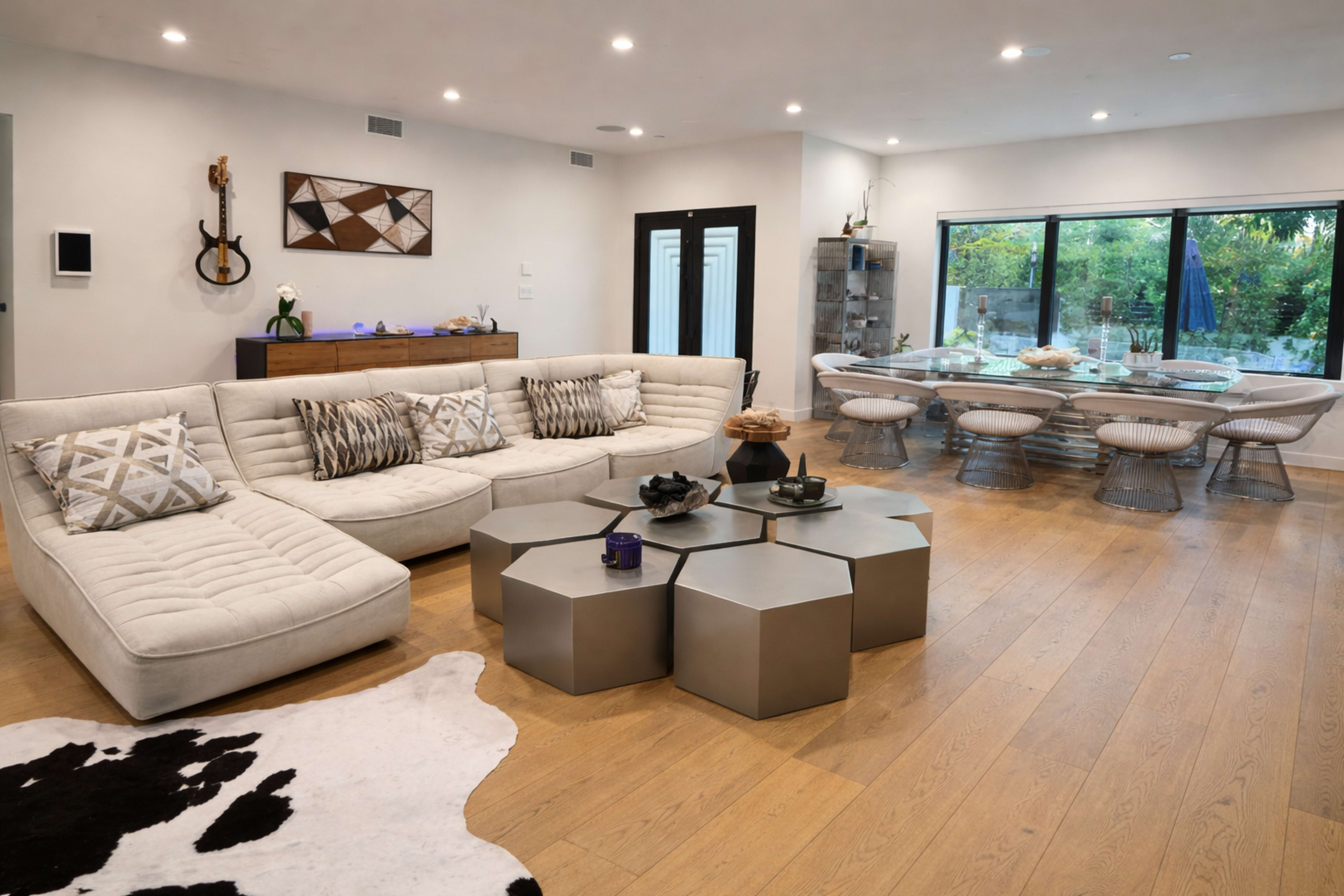 The image shows a modern living room featuring a light-colored sectional sofa, a geometric coffee table, and a dining area with a glass table and metal chairs.