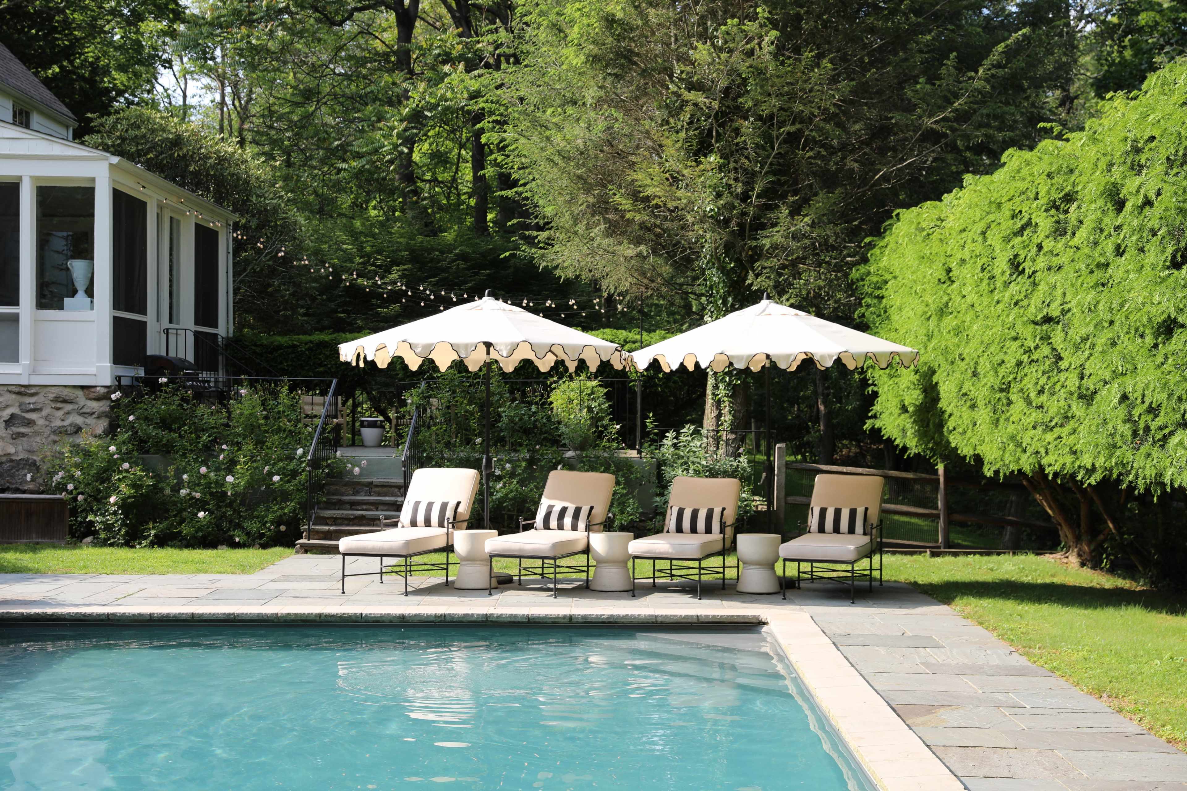 The image shows a serene backyard pool area with four lounge chairs and two umbrellas positioned alongside a clear swimming pool, surrounded by lush greenery.