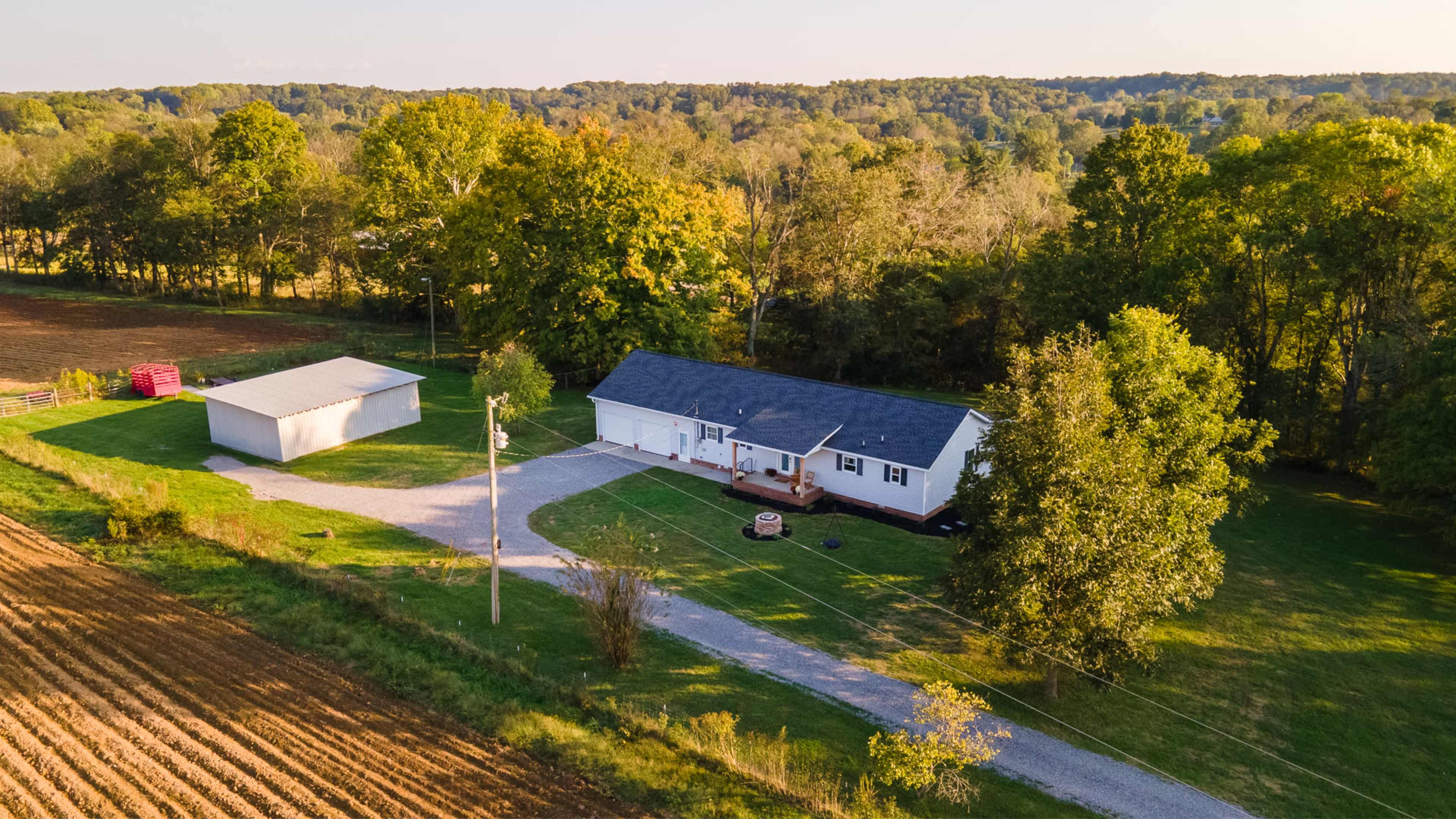 The image shows a single-story white house surrounded by greenery, with a gravel driveway leading to a detached garage, set in a rural landscape.