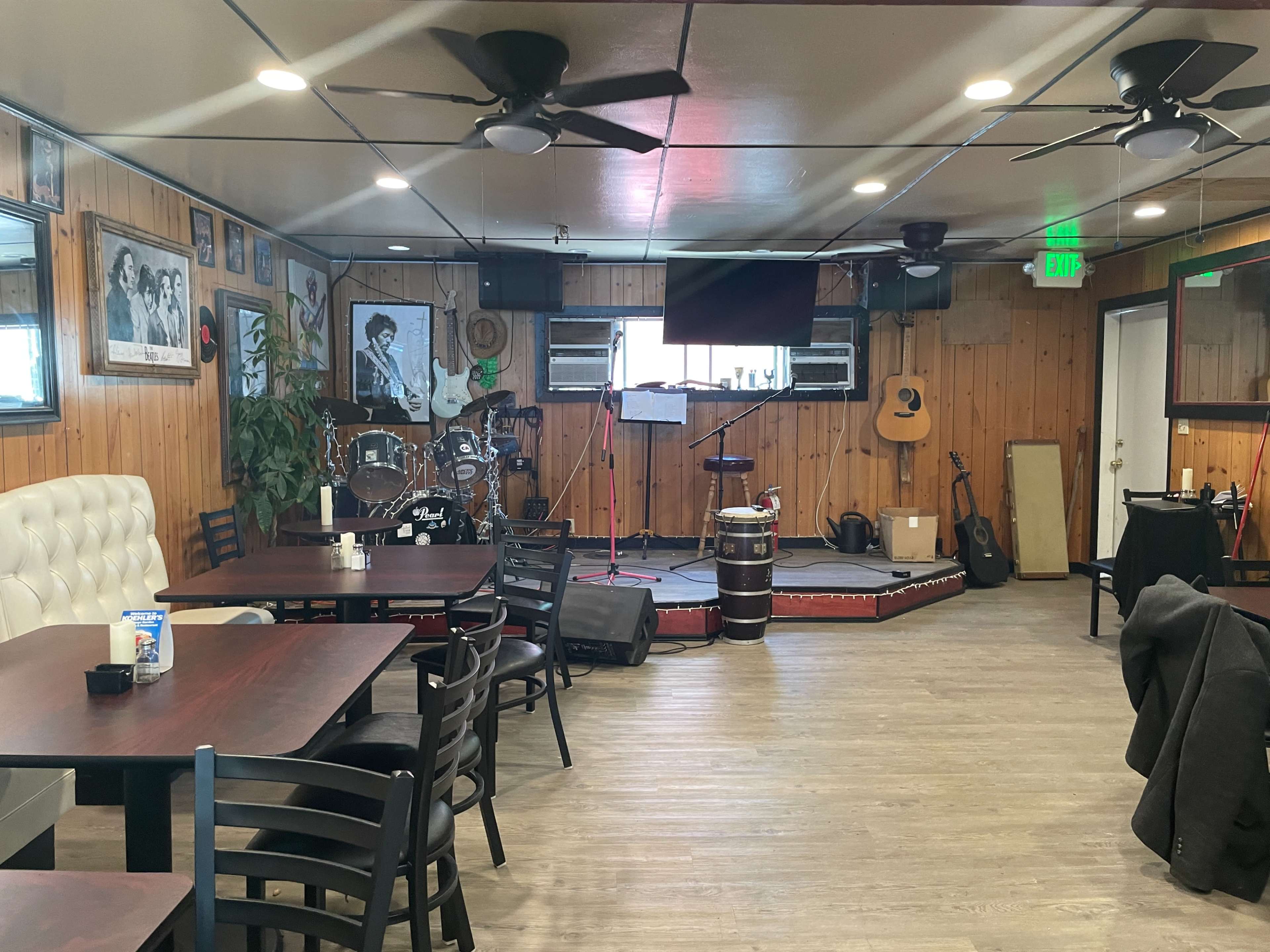 An interior of a restaurant or bar with wooden walls, tables arranged for dining, and a small stage equipped with musical instruments.