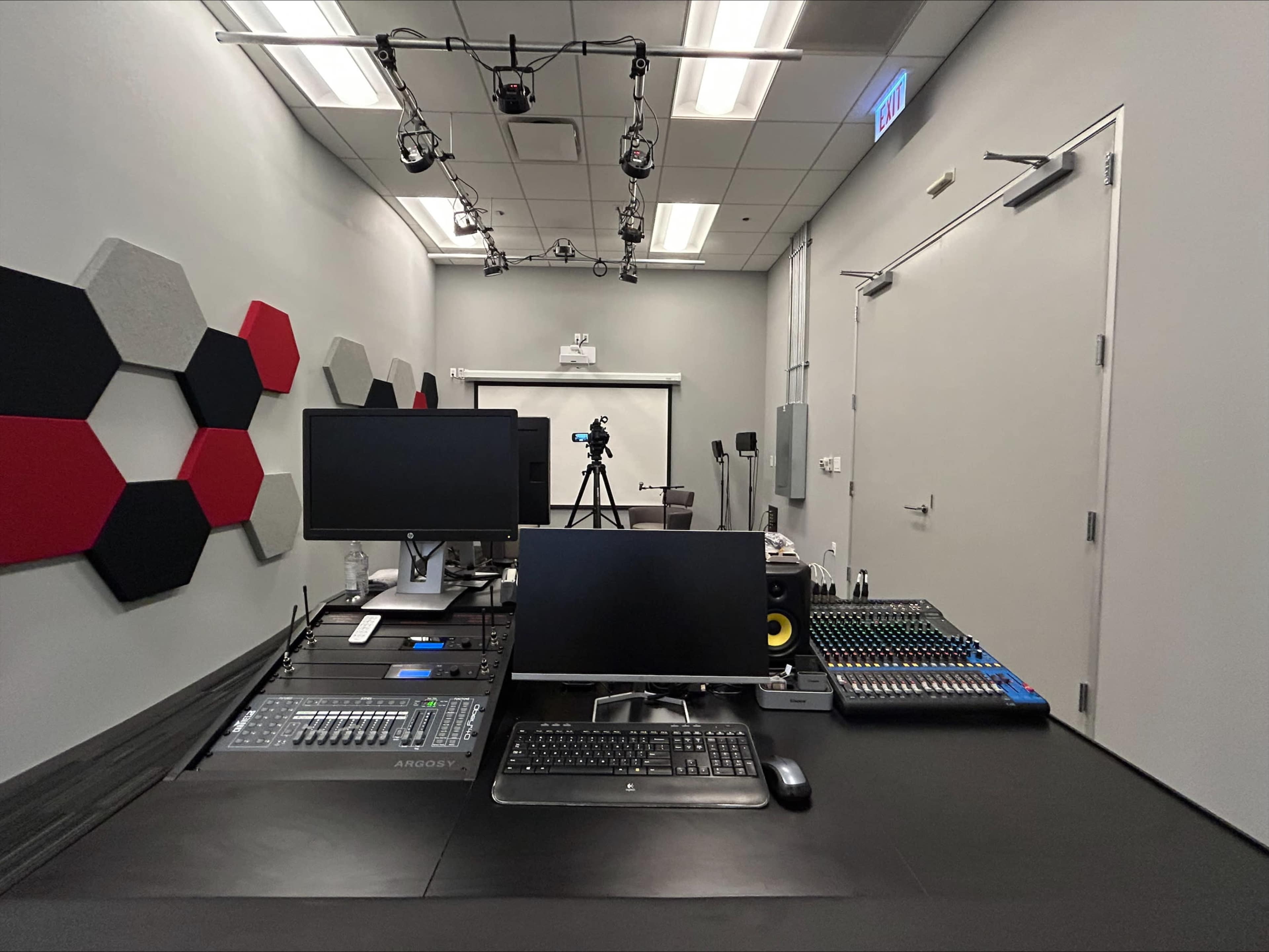 The image shows a recording studio with two computer monitors, a digital audio mixer, a keyboard, and a soundboard set against a backdrop of gray walls and hexagonal acoustic panels.