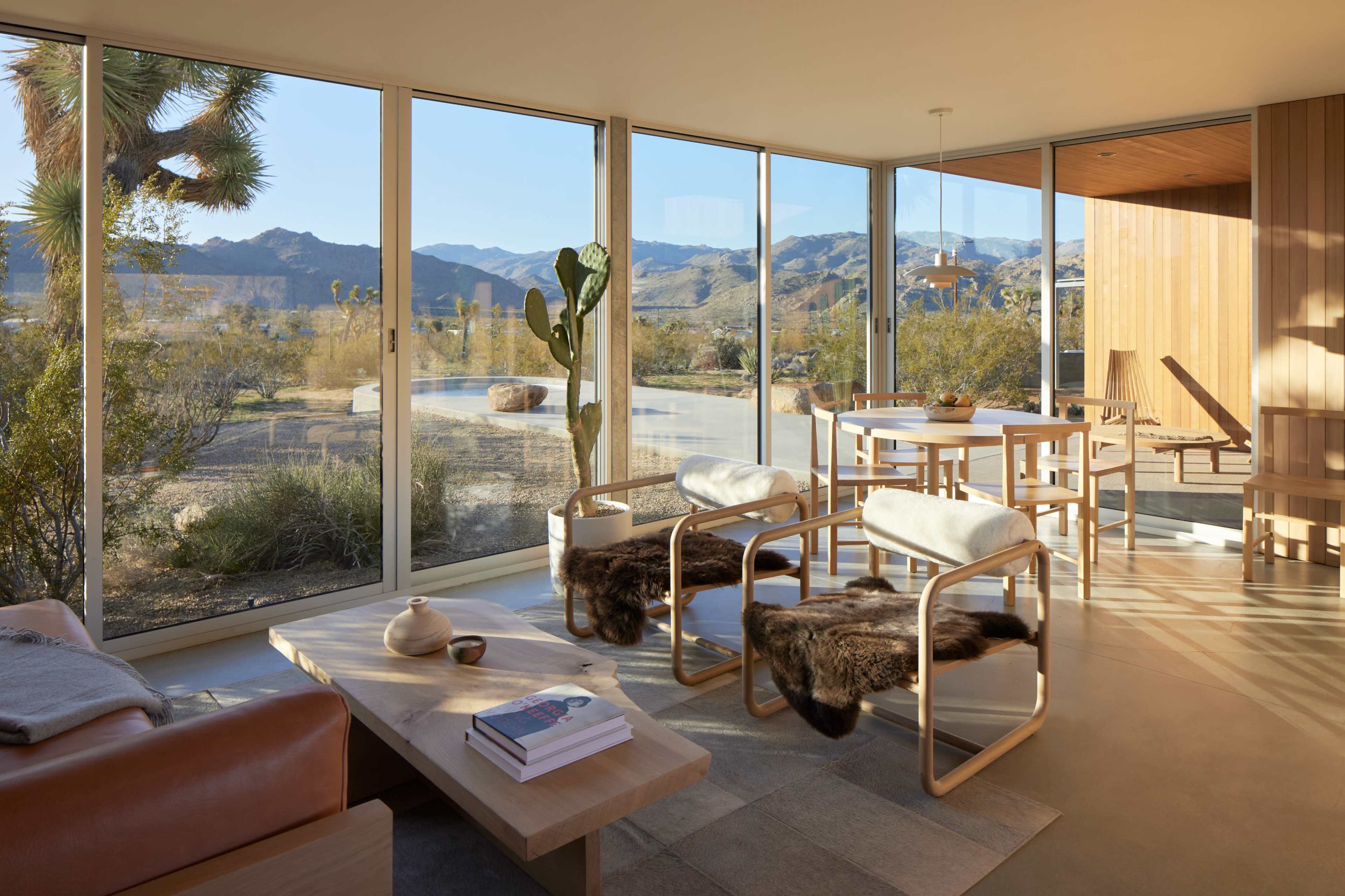The interior of a modern home features large windows that provide a view of mountains and a desert landscape, with minimalist furniture and natural decor elements.