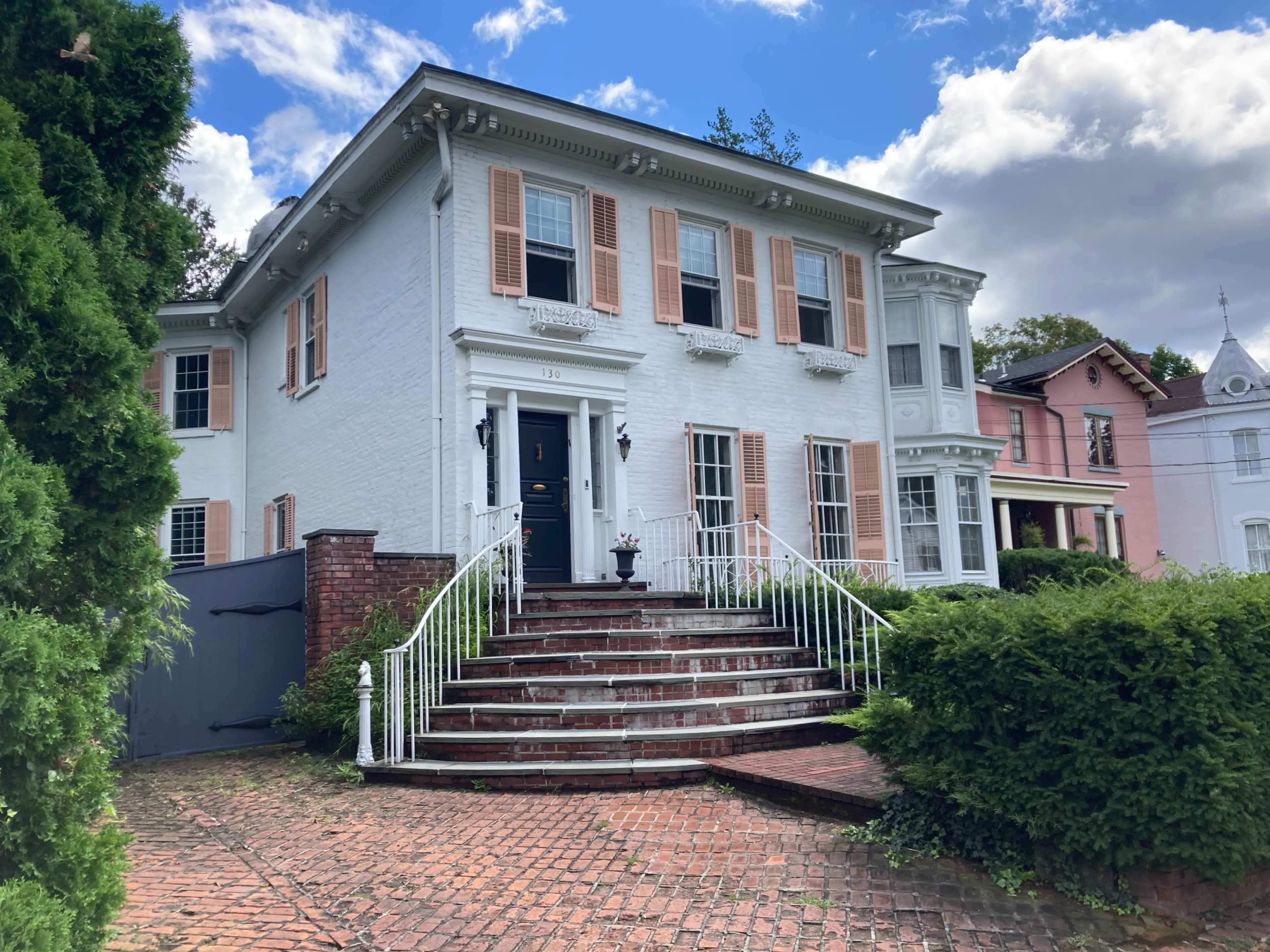 A large white house with brick steps, a black front door, and decorative shutters, flanked by green shrubs and surrounded by other residential buildings.
