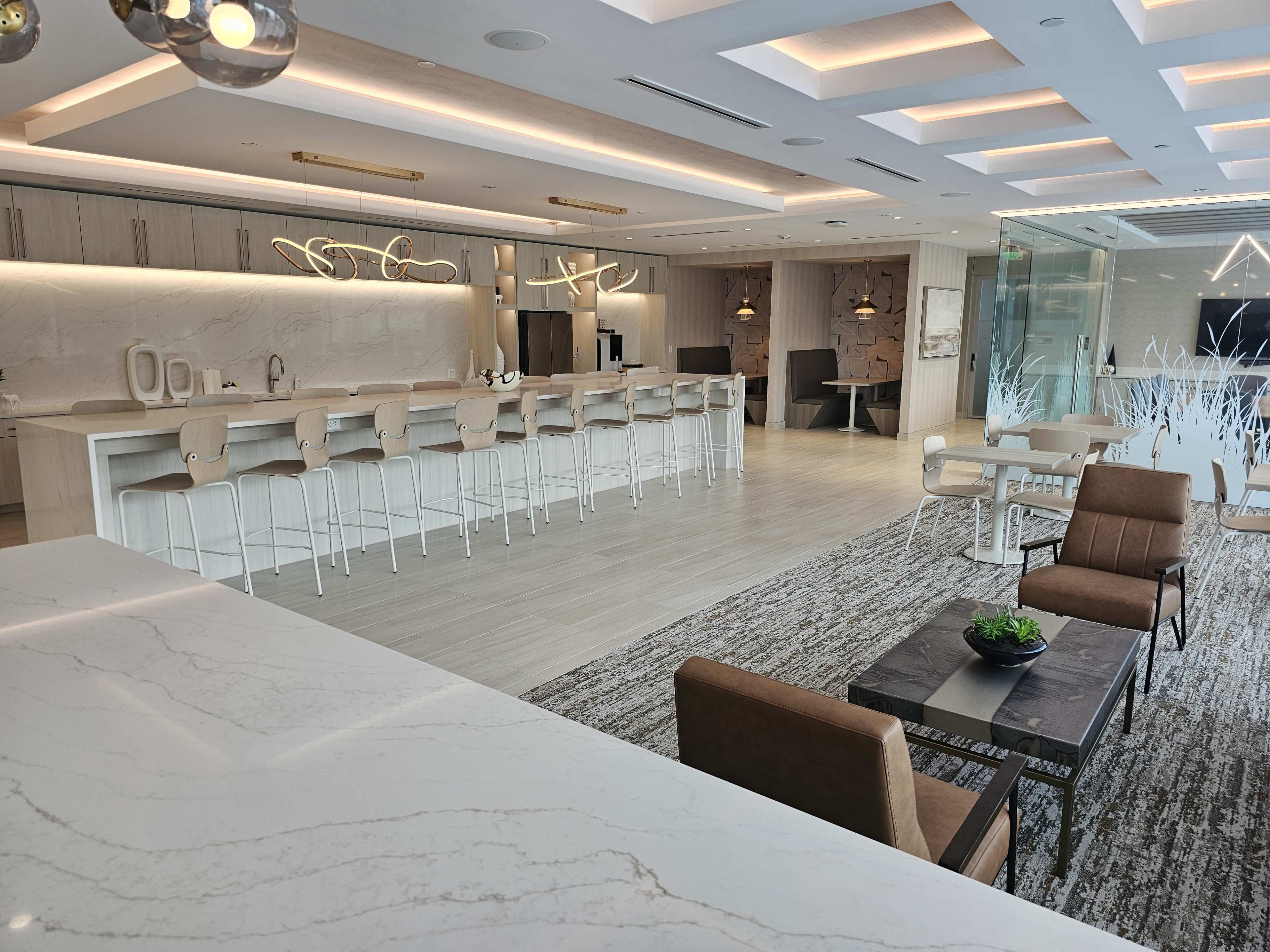 A modern kitchen and lounge area with a long white bar, beige stools, and a seating arrangement featuring two armchairs and a coffee table.