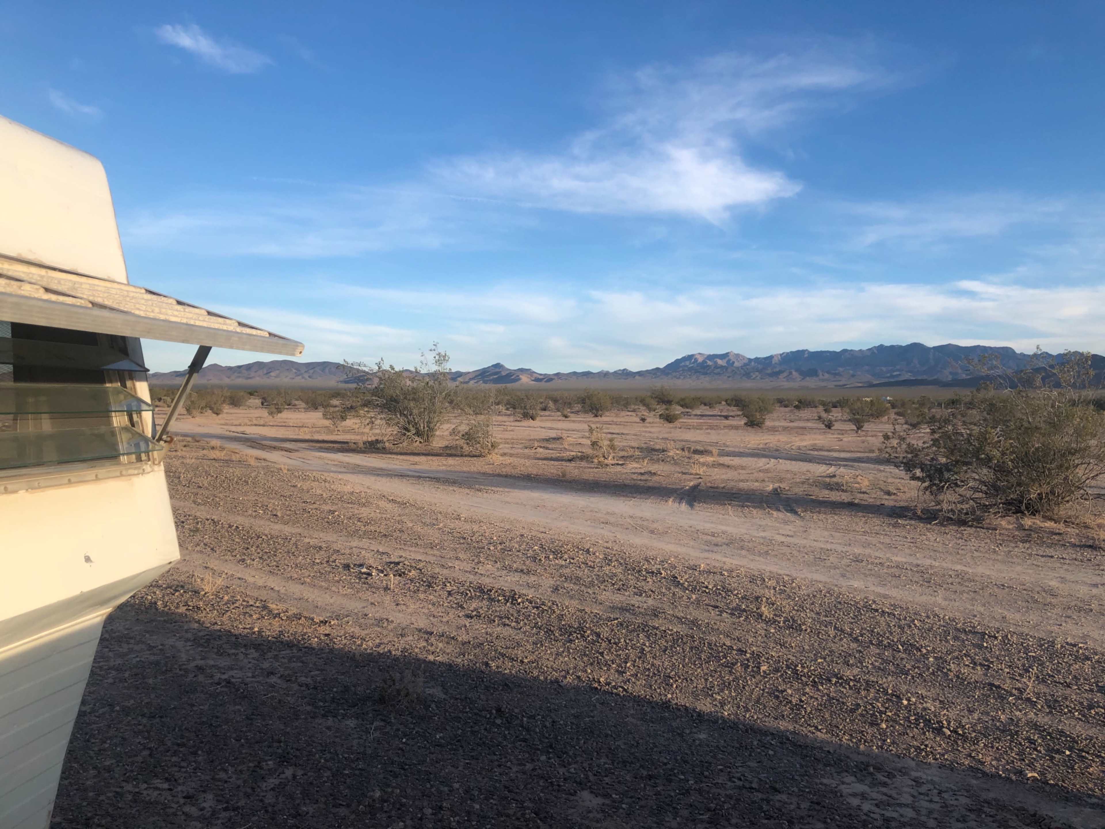 A rugged dirt road leads through a sparse desert landscape with distant mountains under a clear blue sky.