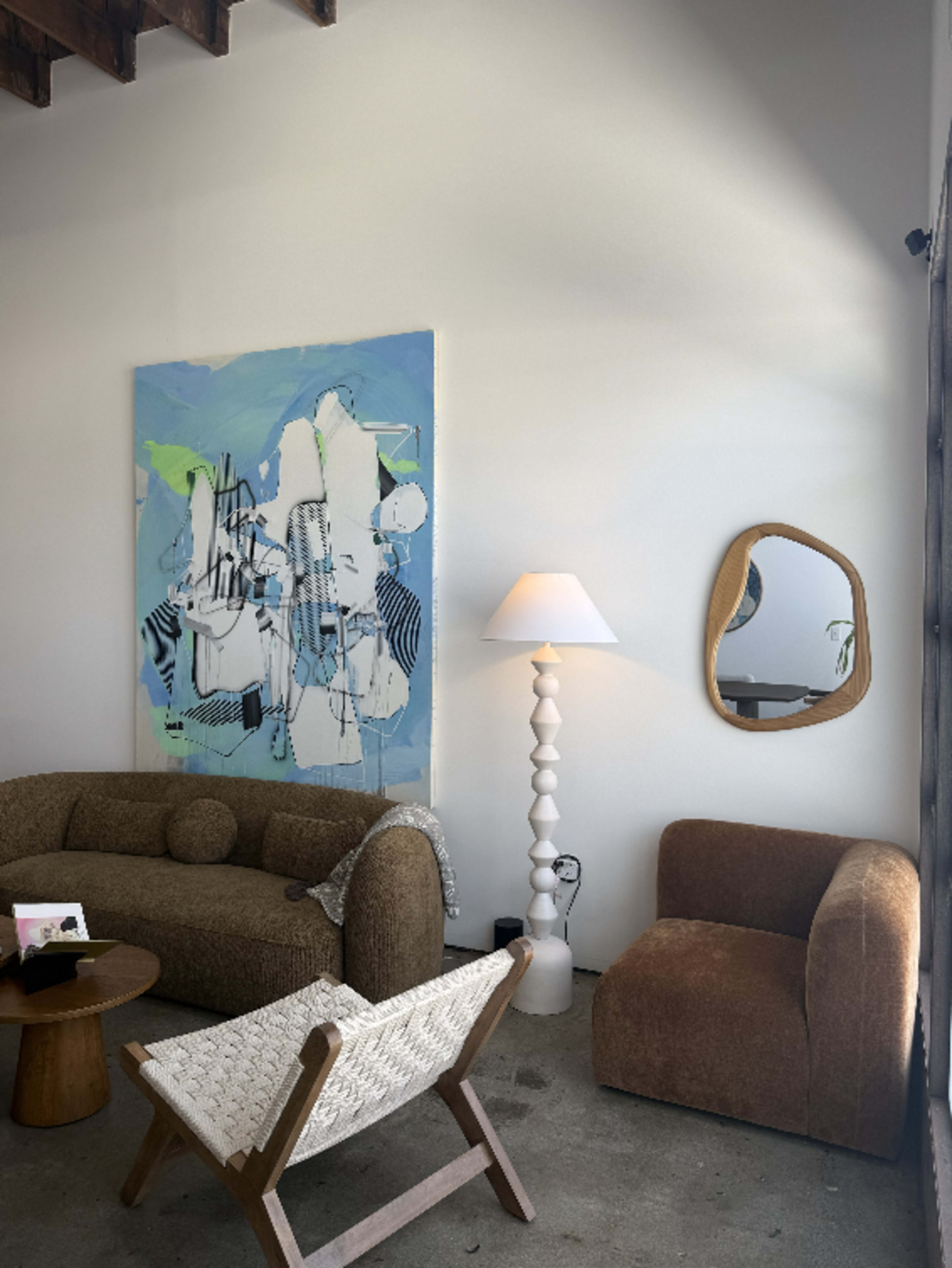 A modern living space features an abstract painting on the wall, a curved brown sofa, a white floor lamp, a small round table, and a mirror with an organic shape.