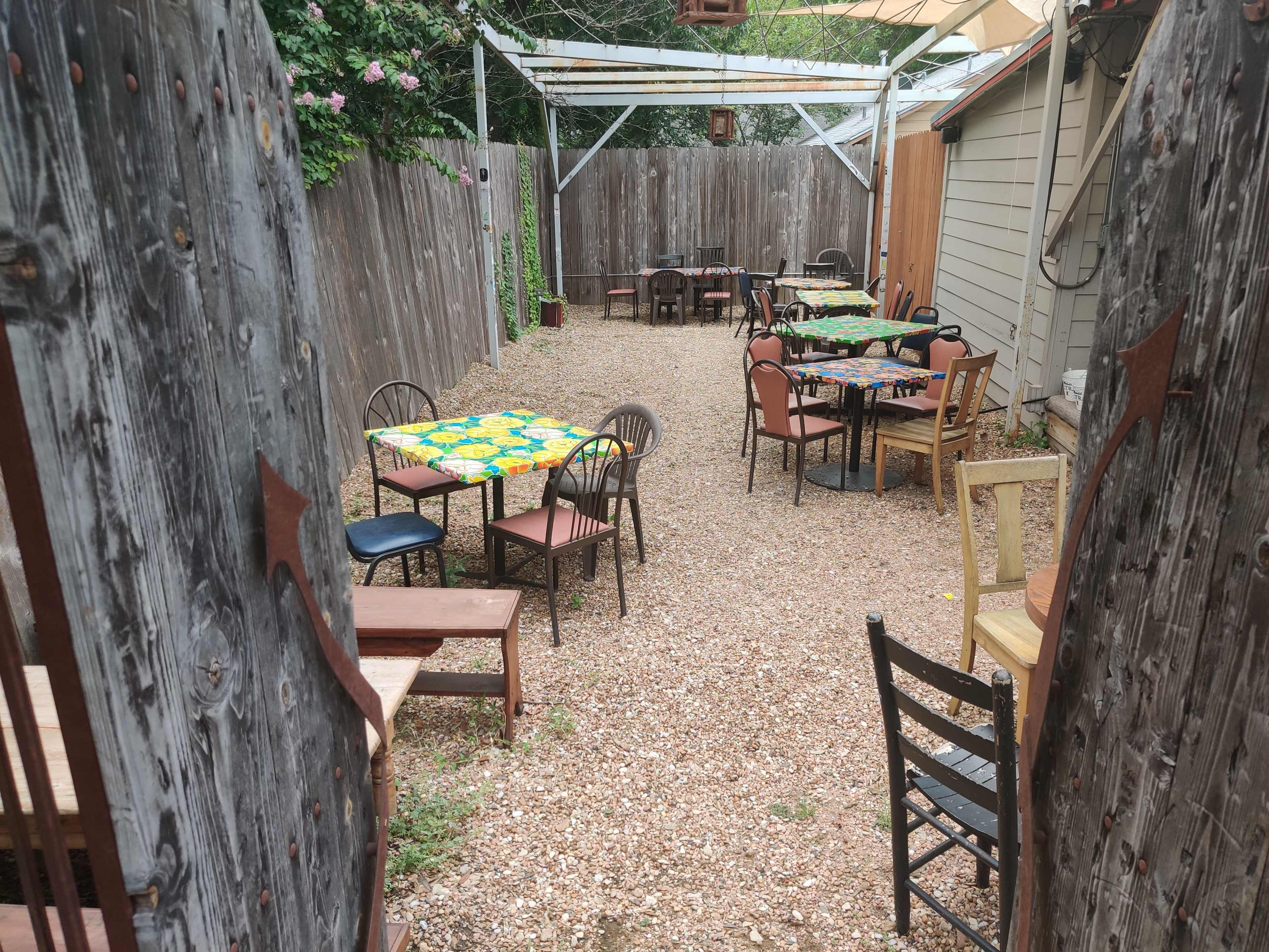 An outdoor dining area with scattered tables and chairs set on a gravel surface, surrounded by wooden fencing.