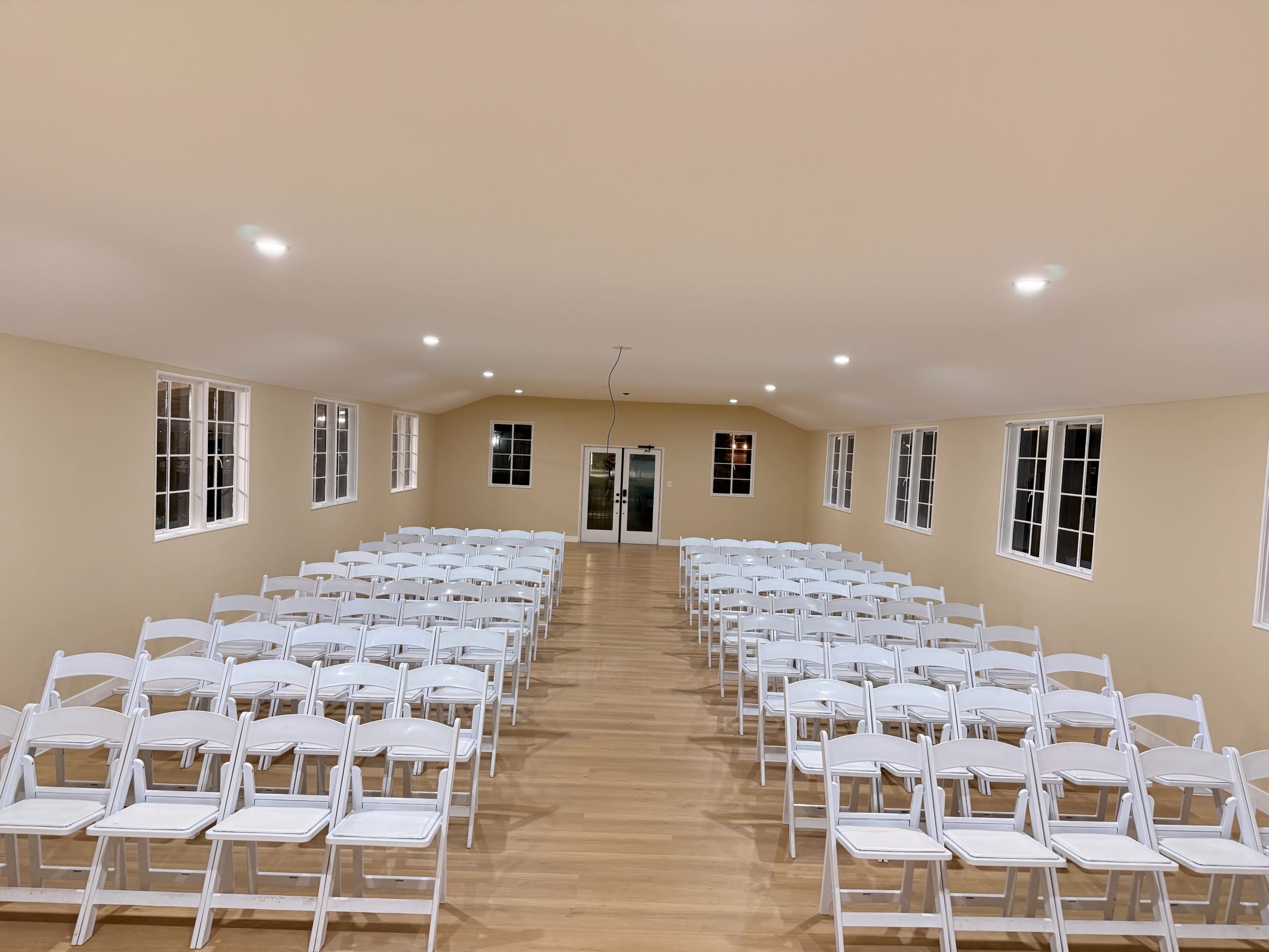 The image shows a large, empty room with rows of white chairs arranged facing a door at the far end.