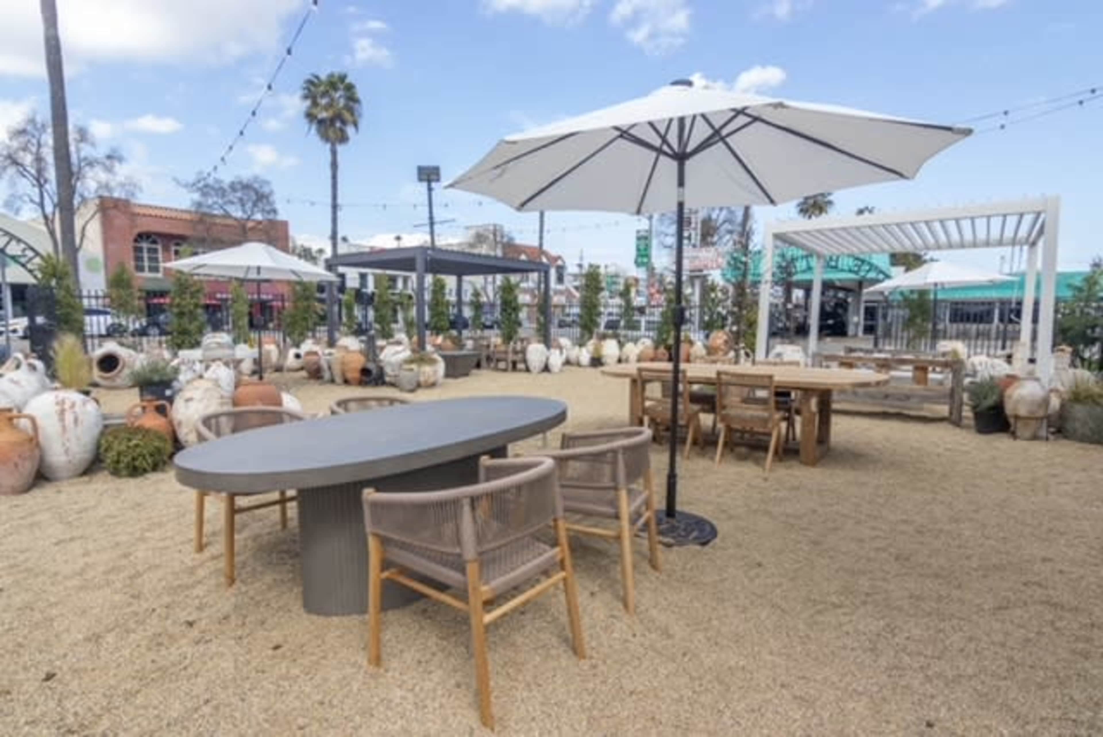 An outdoor seating area with several tables, chairs, and large umbrellas, surrounded by decorative pottery and greenery on a gravel surface.
