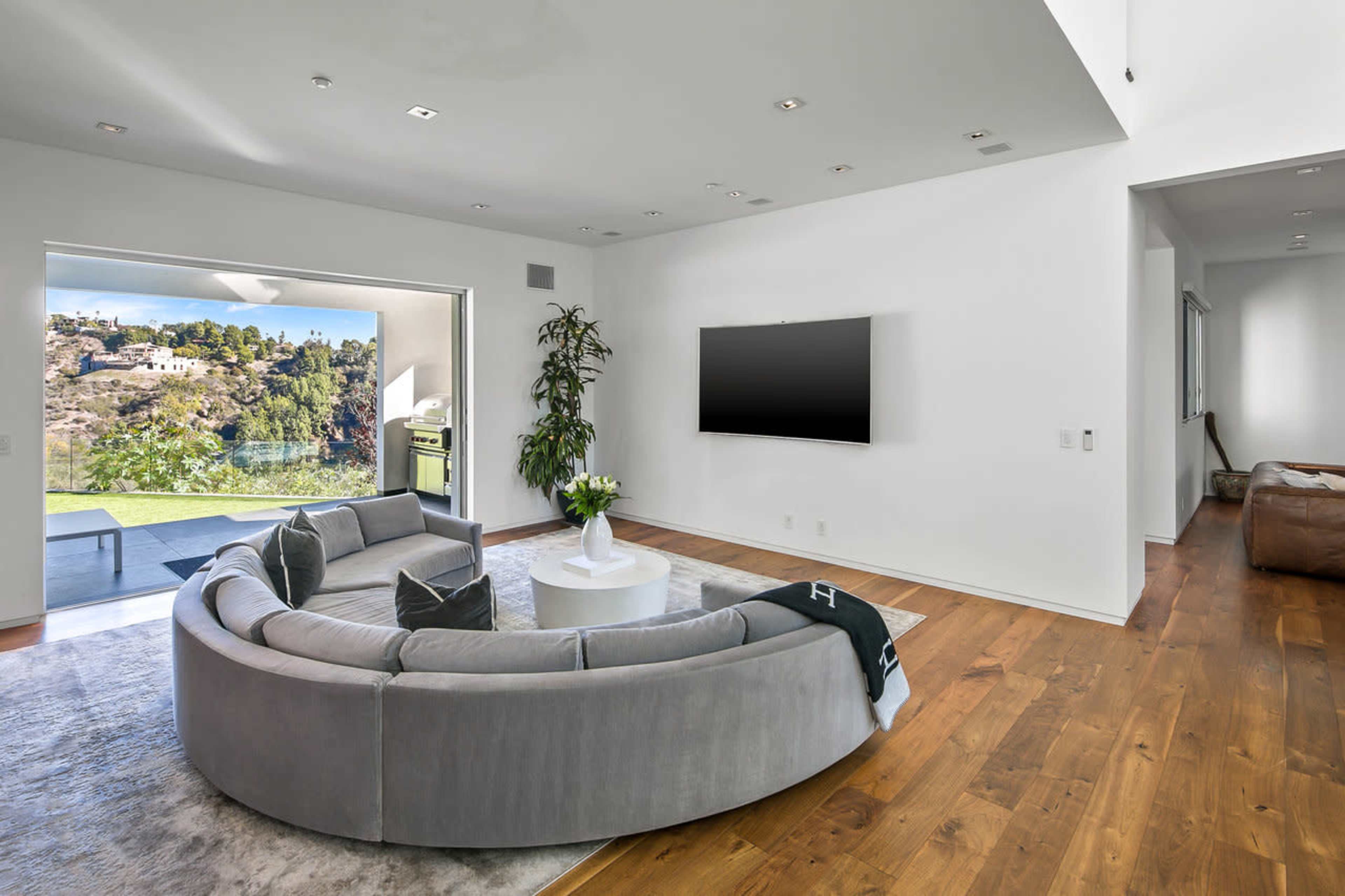 A modern living room features a curved gray sofa, a round white coffee table, and large windows overlooking a scenic view.