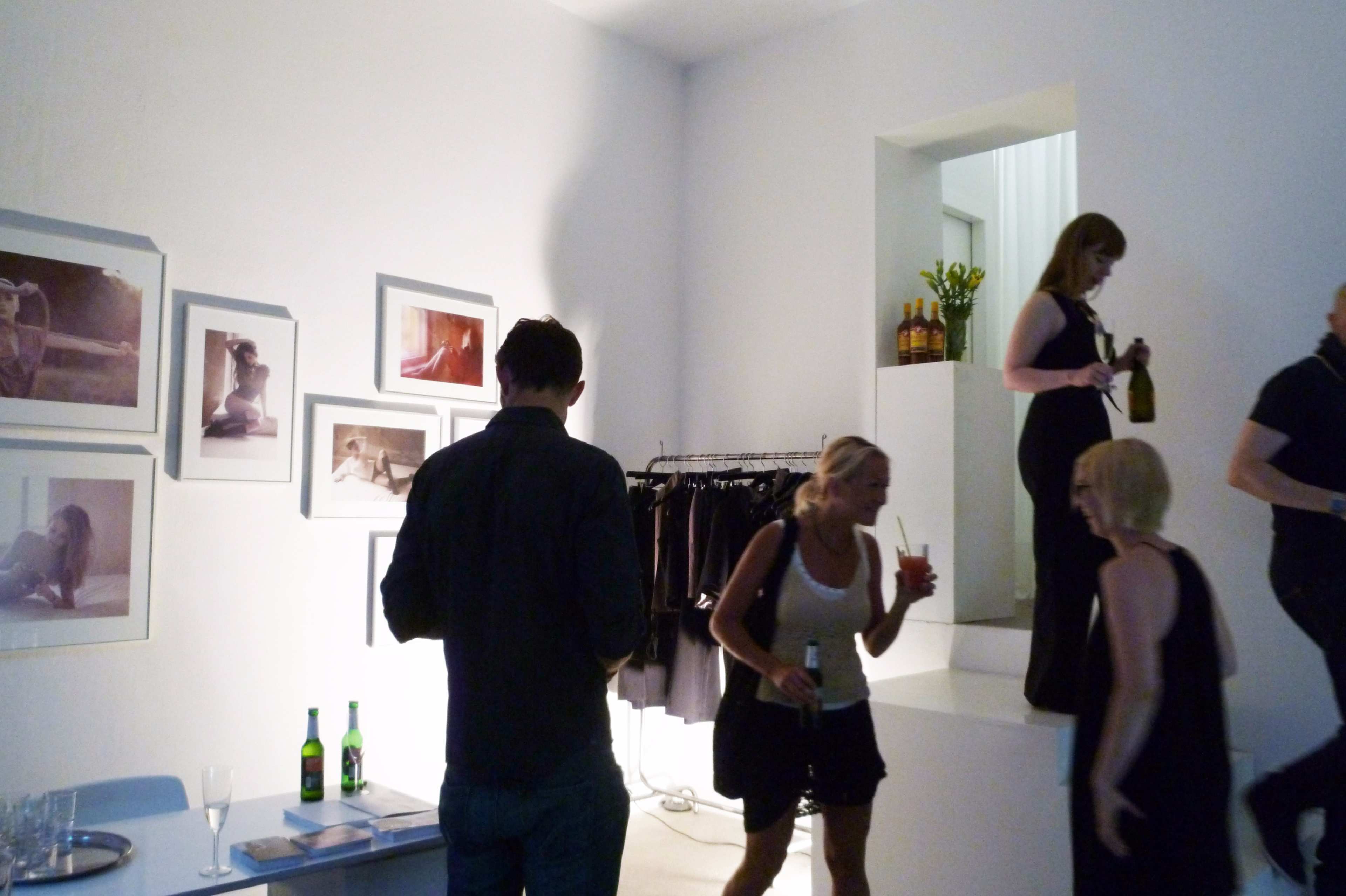 A group of people mingles in a minimalistic space adorned with framed photographs and clothing racks, with some individuals holding drinks.
