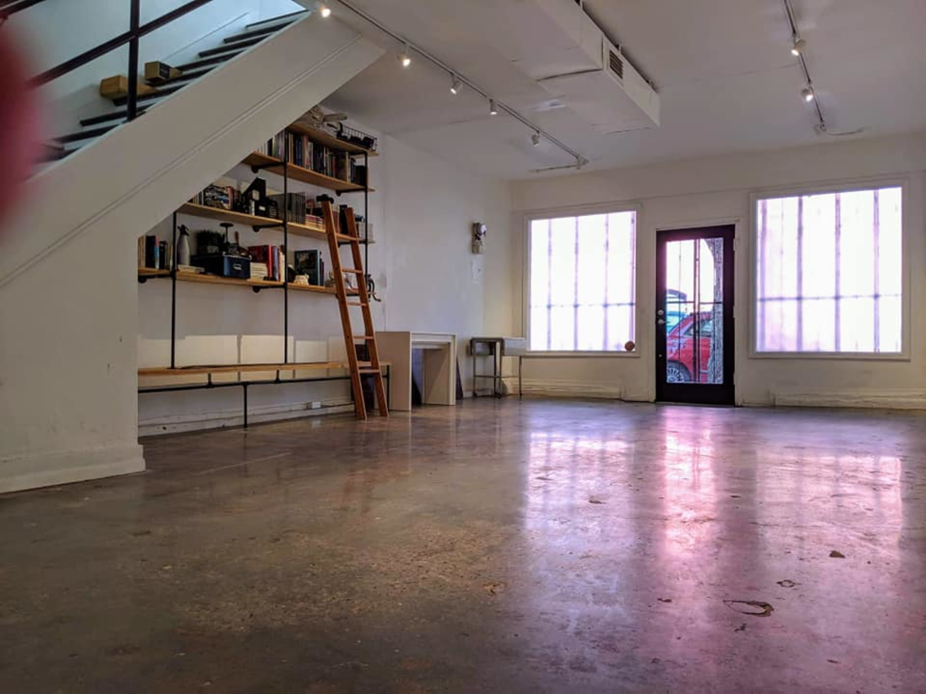 The image shows a spacious room with a polished concrete floor, a wooden ladder against a shelf filled with items, and large windows at the front.