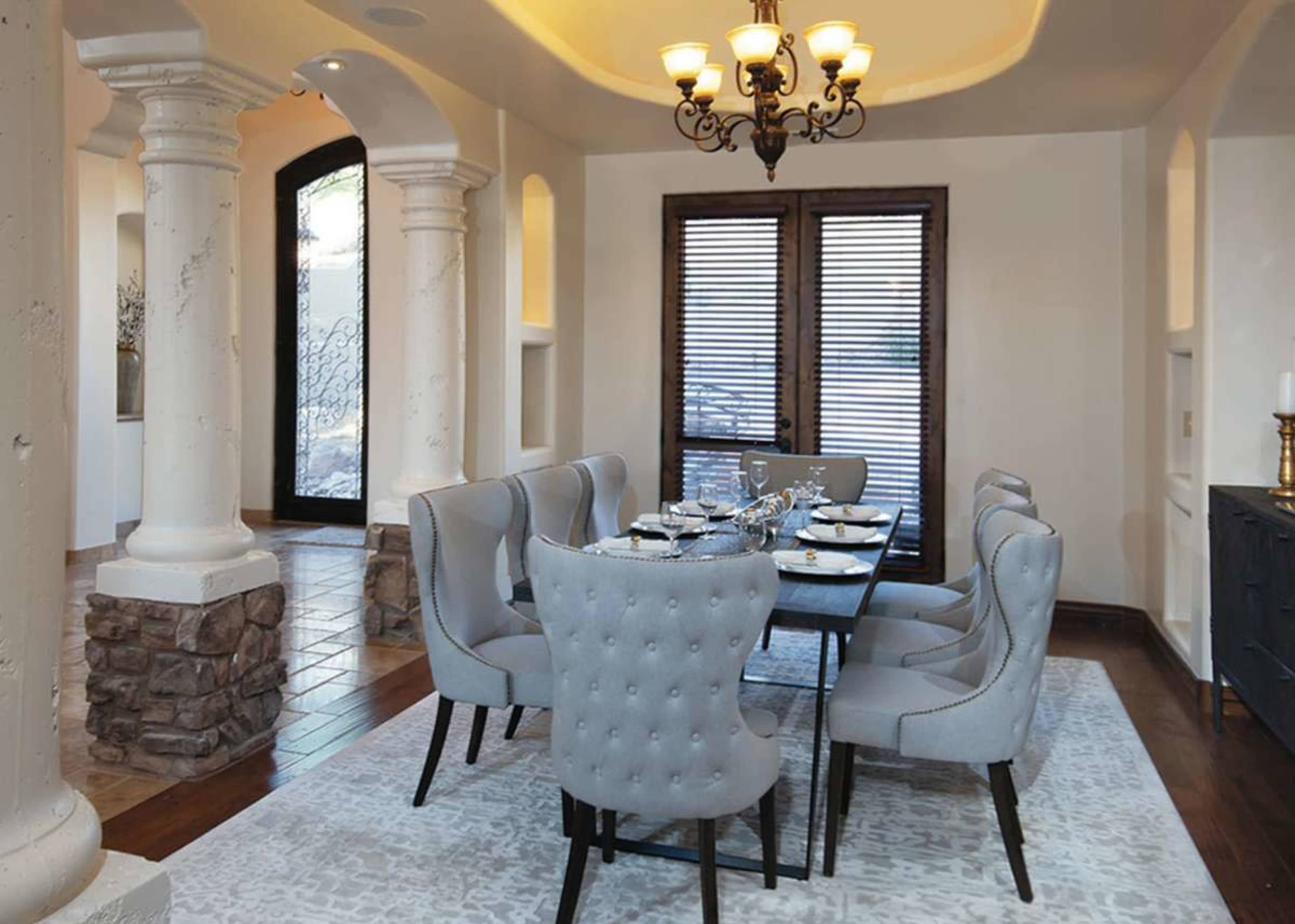 A dining room features a rectangular table surrounded by six upholstered chairs, with stone columns and large windows in the background.