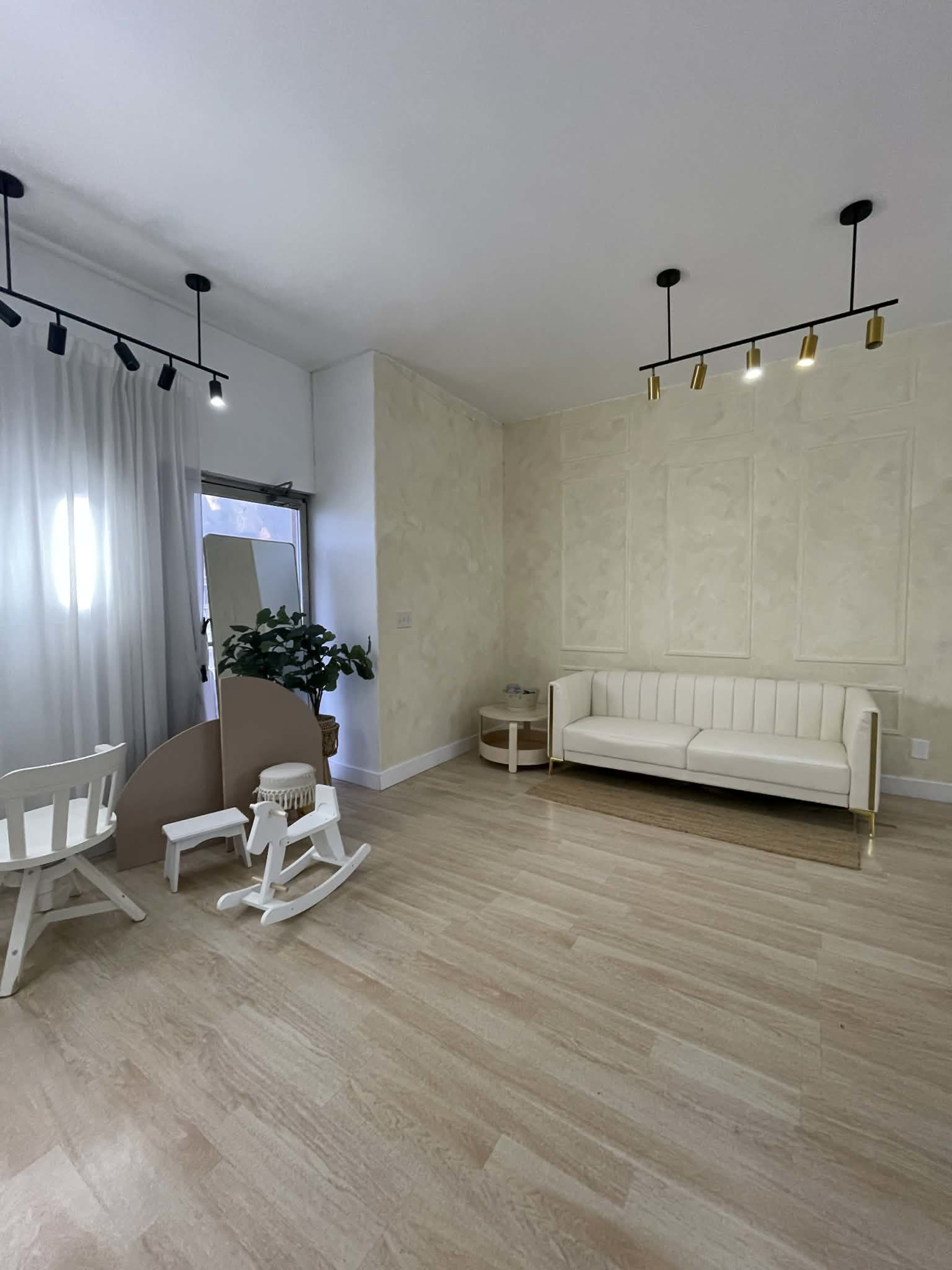 The image shows a minimalistic, light-colored room featuring a white couch, a small wooden rocking chair, and a potted plant near a window.