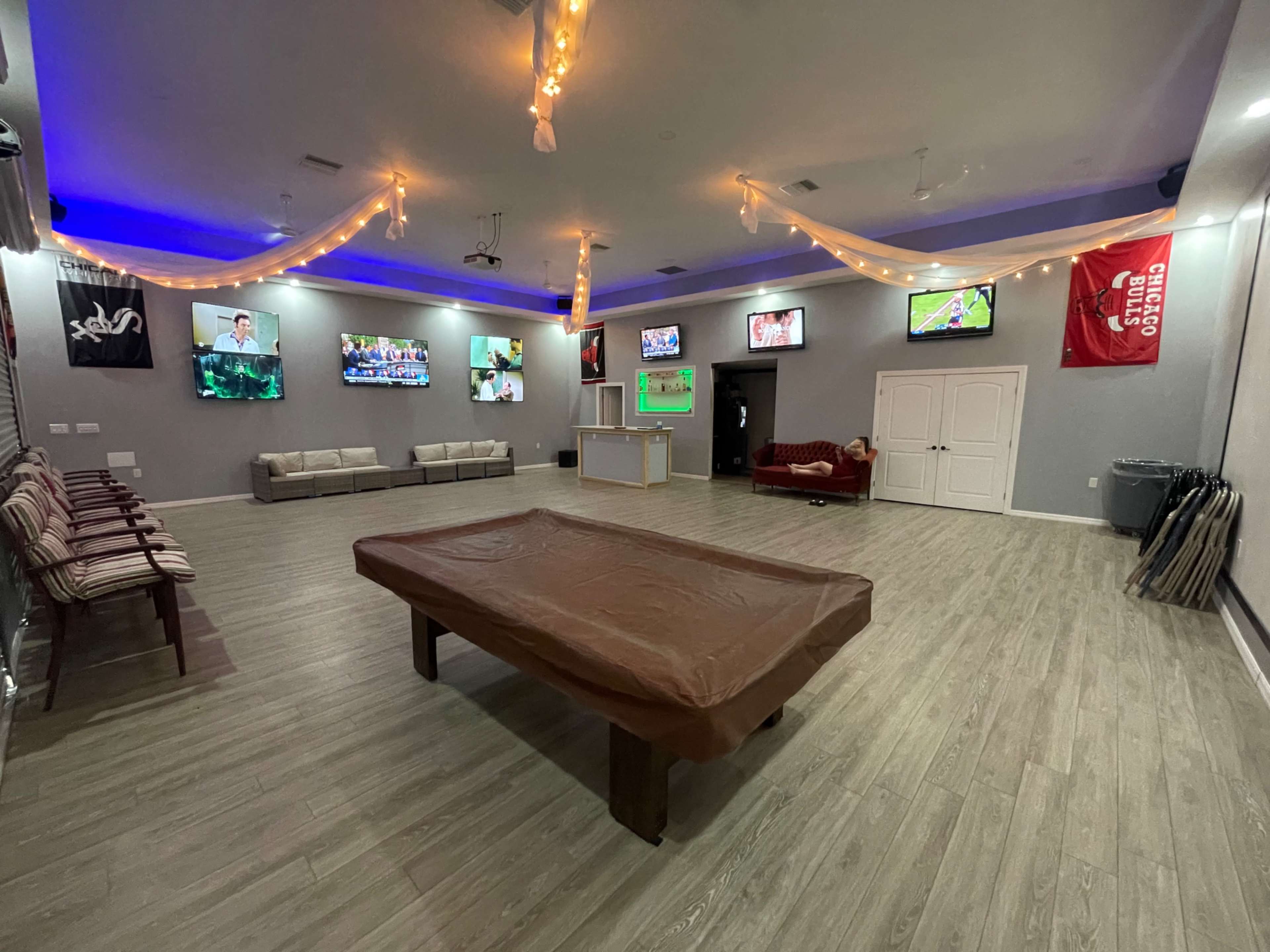 The image shows a spacious recreation room with multiple televisions displaying sports, a pool table covered with a protective cover, and seating arranged along the walls.