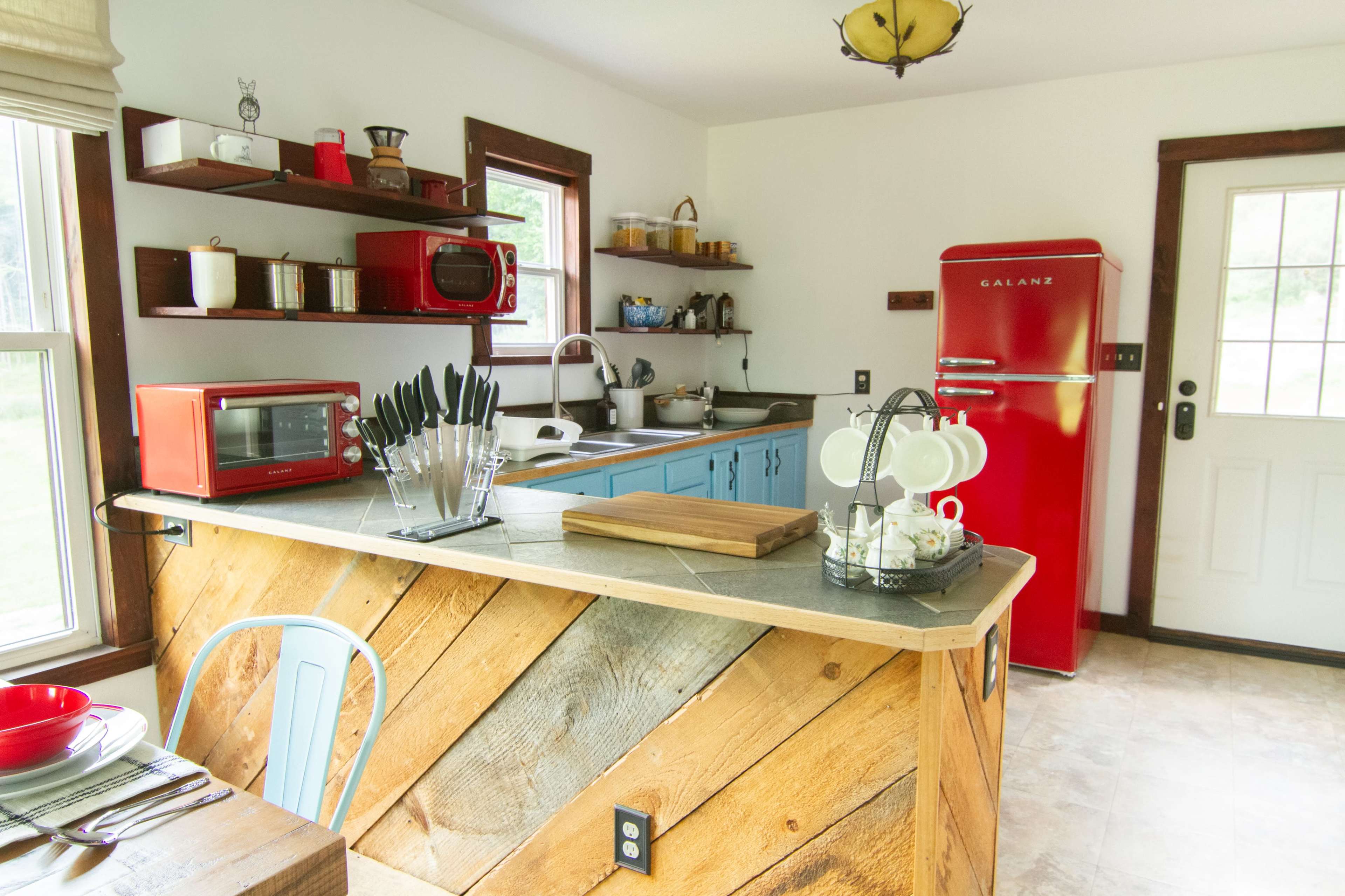 The image shows a bright kitchen with wooden countertops, blue cabinets, a red refrigerator, and a variety of kitchen appliances and utensils on display.