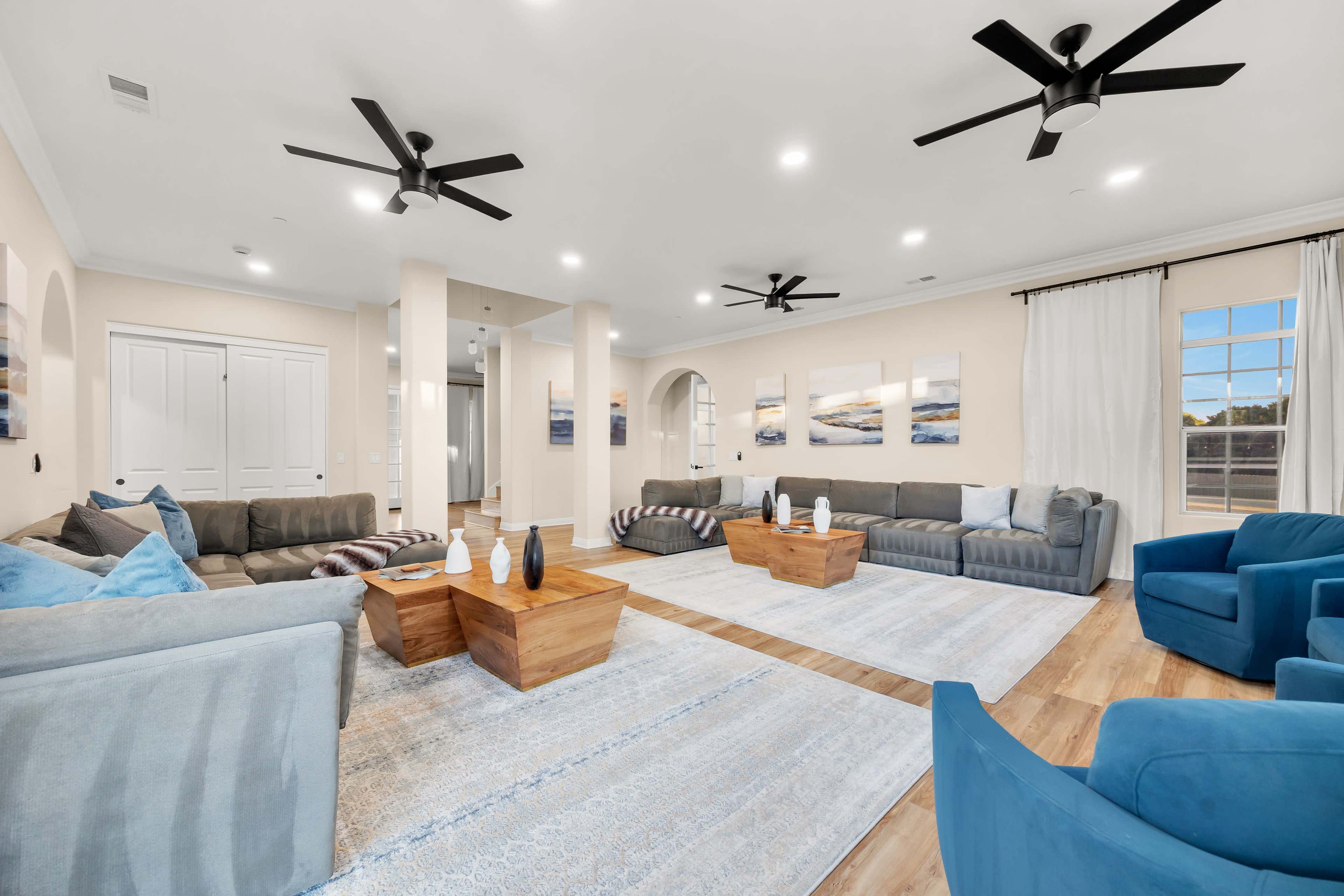 The image shows a spacious living room with multiple seating areas, featuring gray sofas, blue accent chairs, wooden coffee tables, and large windows.