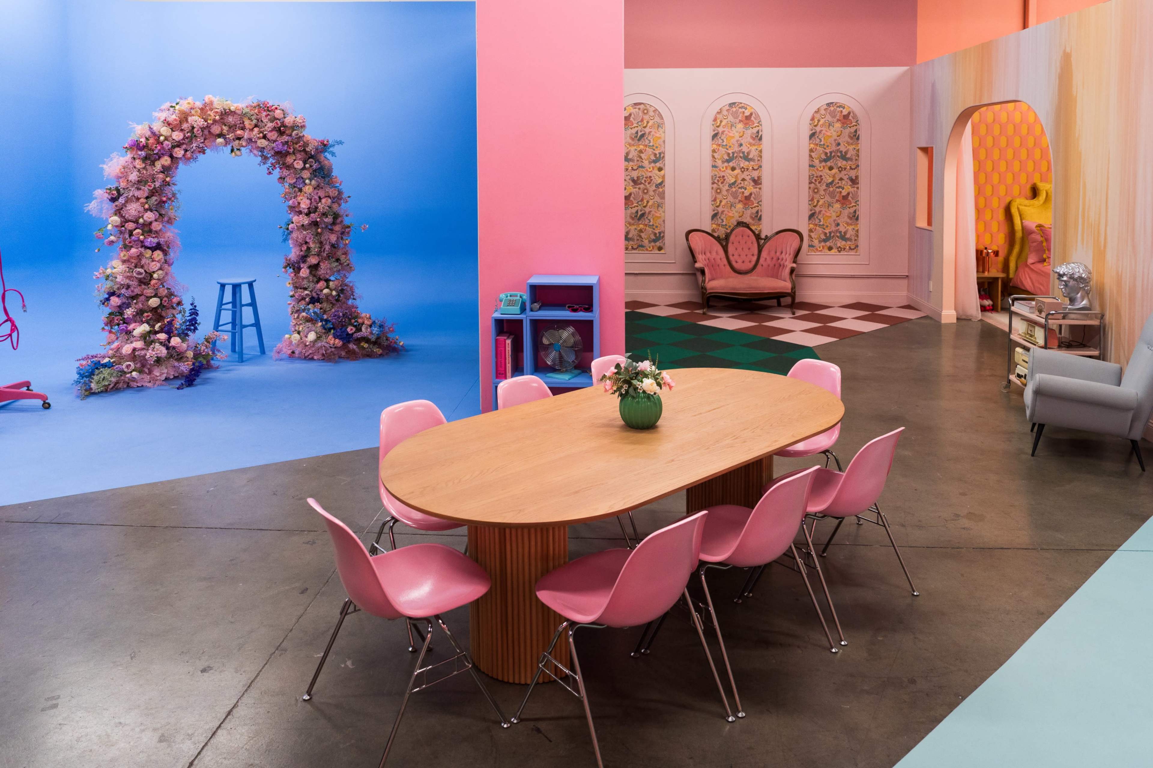 The scene features a brightly colored room with a large wooden dining table surrounded by pink chairs, set against a backdrop of floral arches, patterned walls, and a mix of furniture styles.