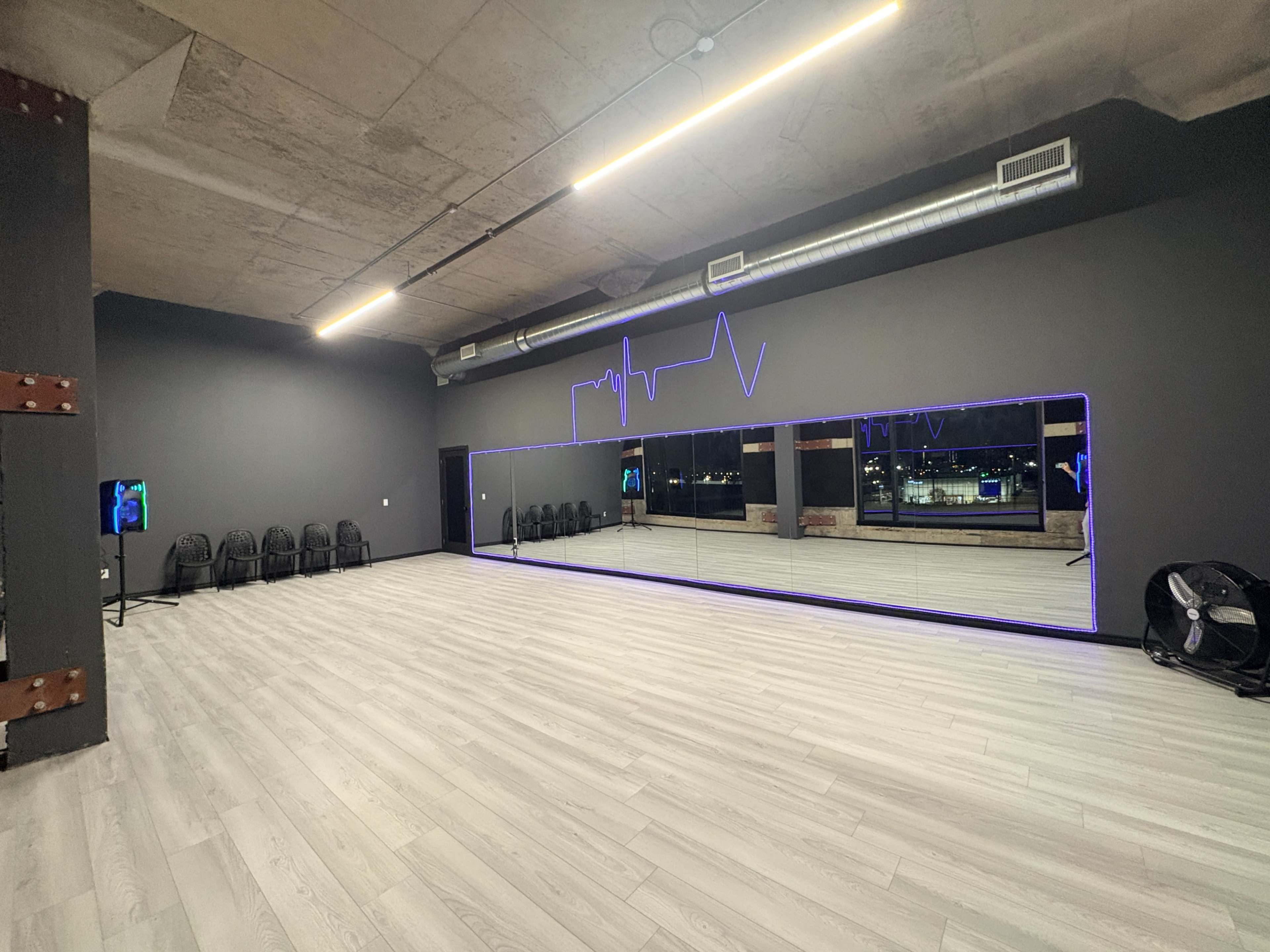 The image shows a spacious, modern studio with a large mirror along one wall, accented by purple LED lights, and empty chairs lined up against another wall.