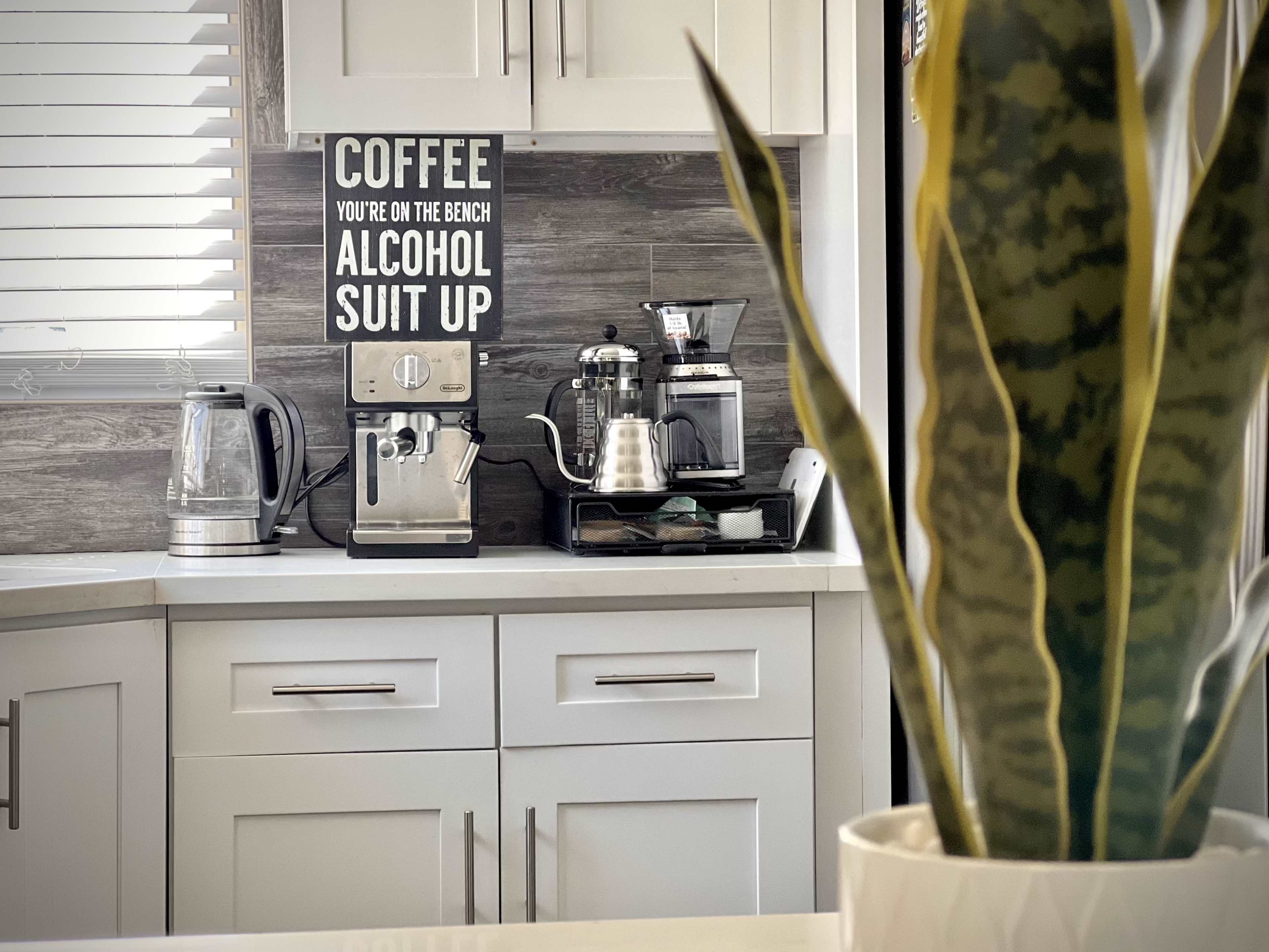 A kitchen counter features a sign that reads "COFFEE YOU'RE ON THE BENCH ALCOHOL SUIT UP," alongside various coffee-making appliances and a potted plant.