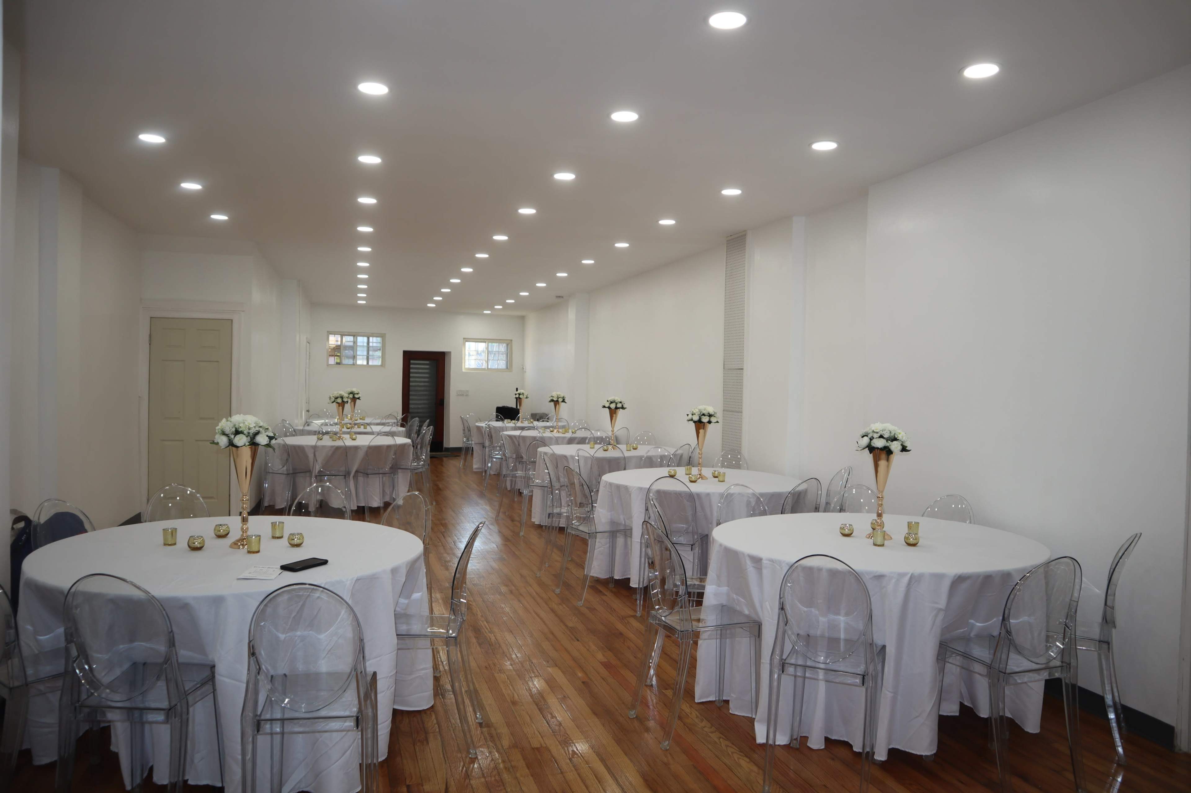 A well-lit banquet hall features several round tables with white tablecloths and clear plastic chairs arranged along a polished wooden floor.