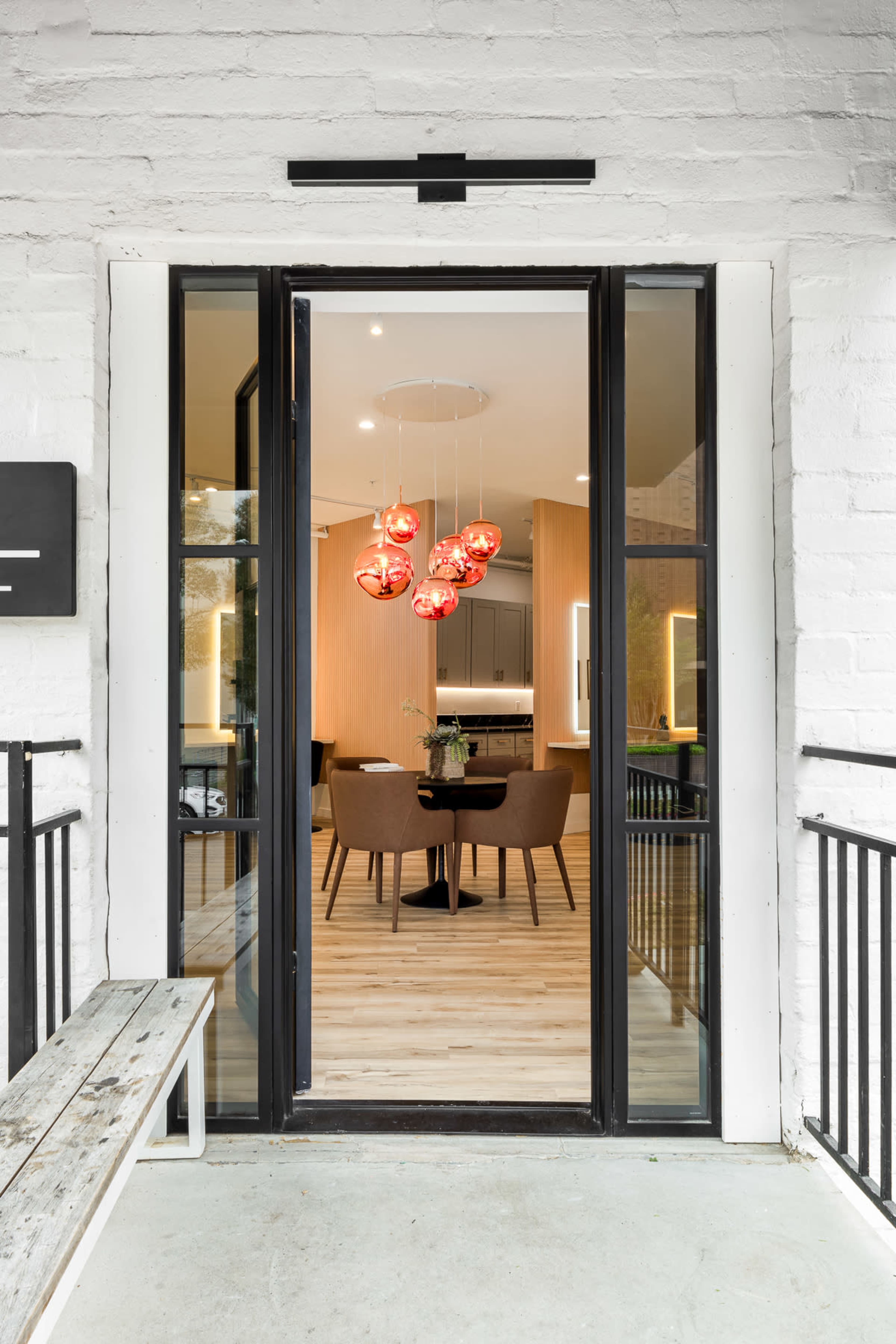 The image shows a view through a doorway with black-framed glass, revealing a modern interior featuring a dining area with pendant lights and wooden flooring.