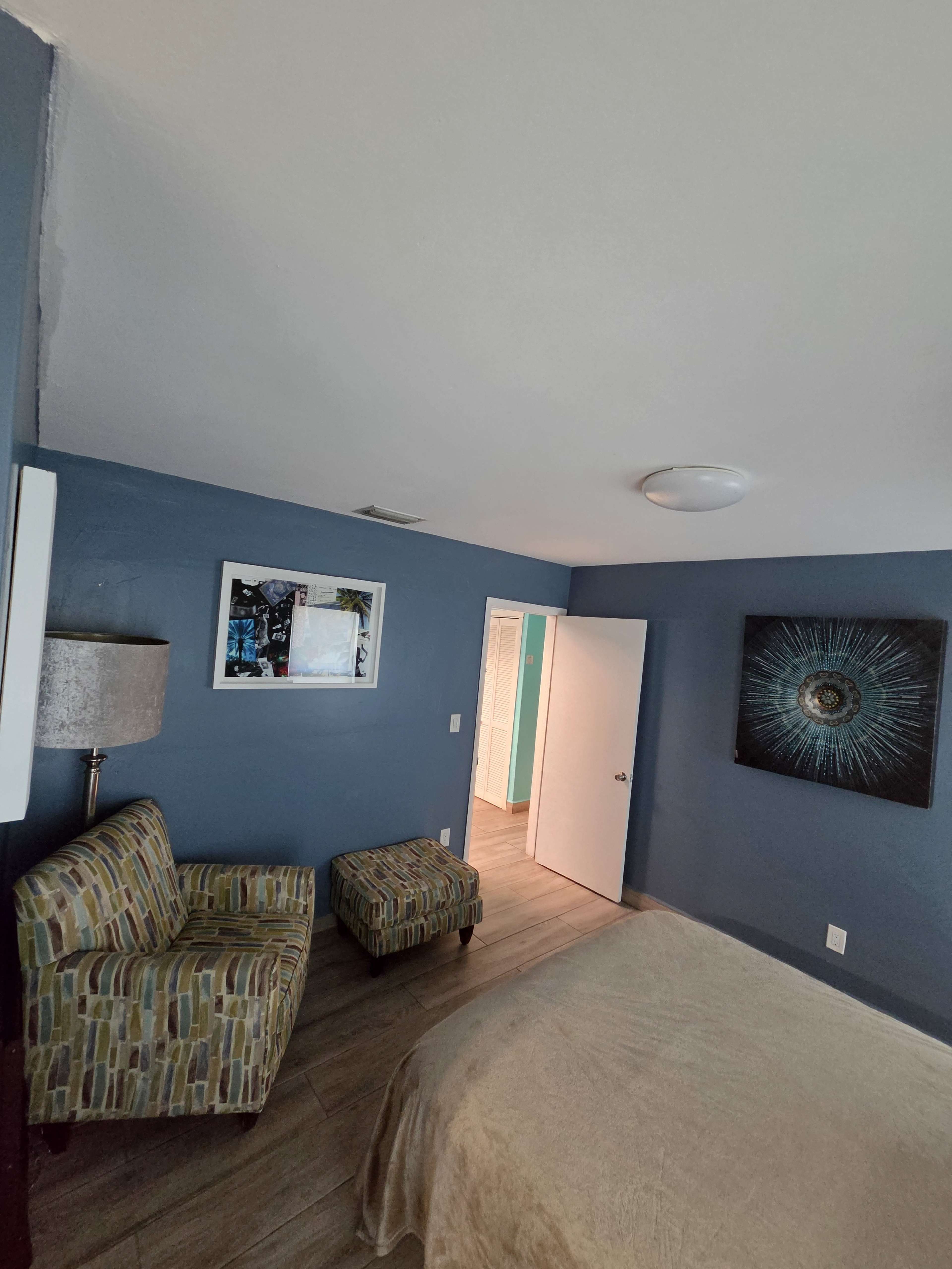 The image shows a bedroom with a blue wall, a bed covered with a beige blanket, a patterned chair and ottoman, and an open door leading to a brightly lit hallway.