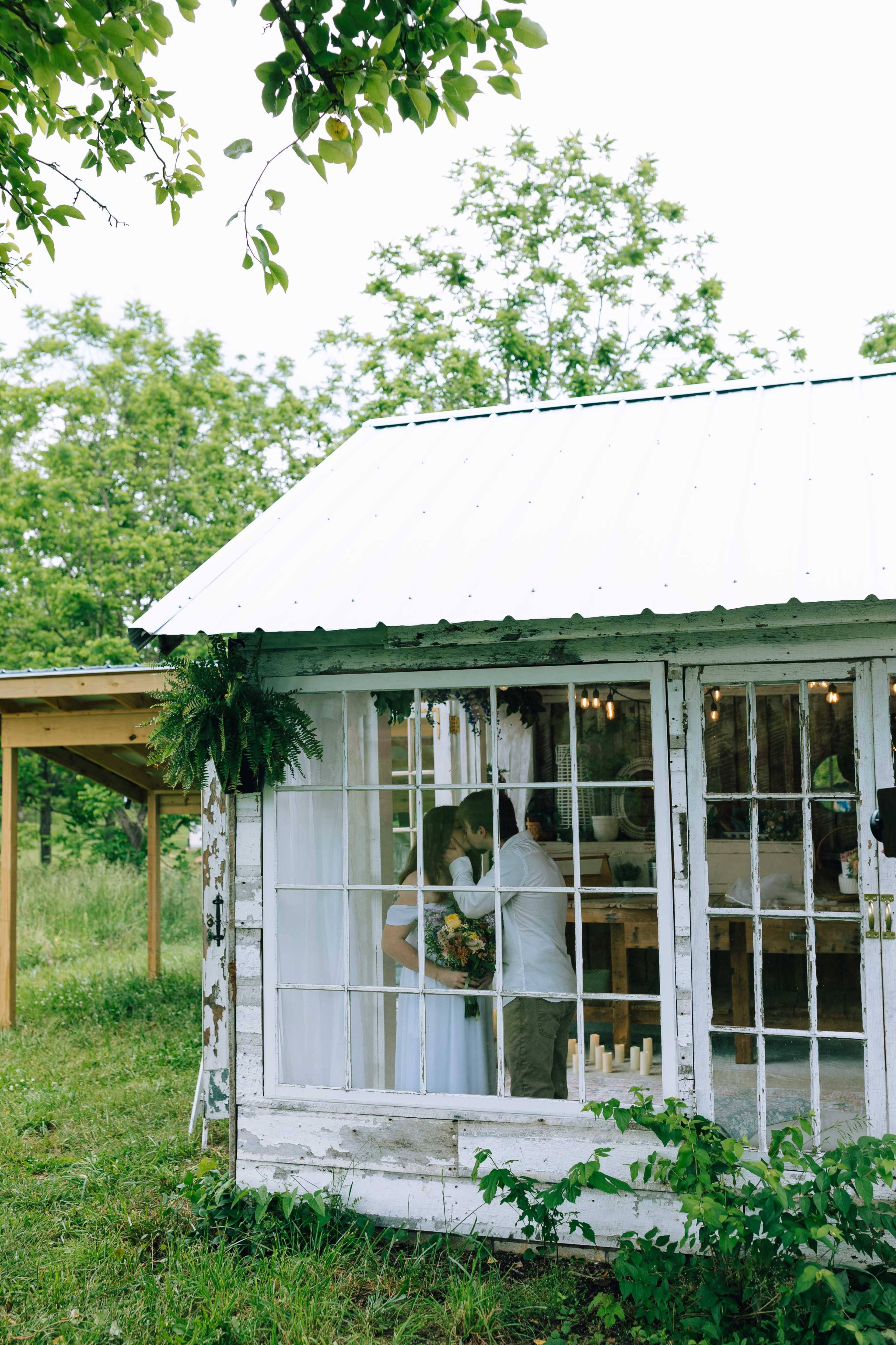 A couple embraces inside a rustic white cabin with large windows, surrounded by greenery.
