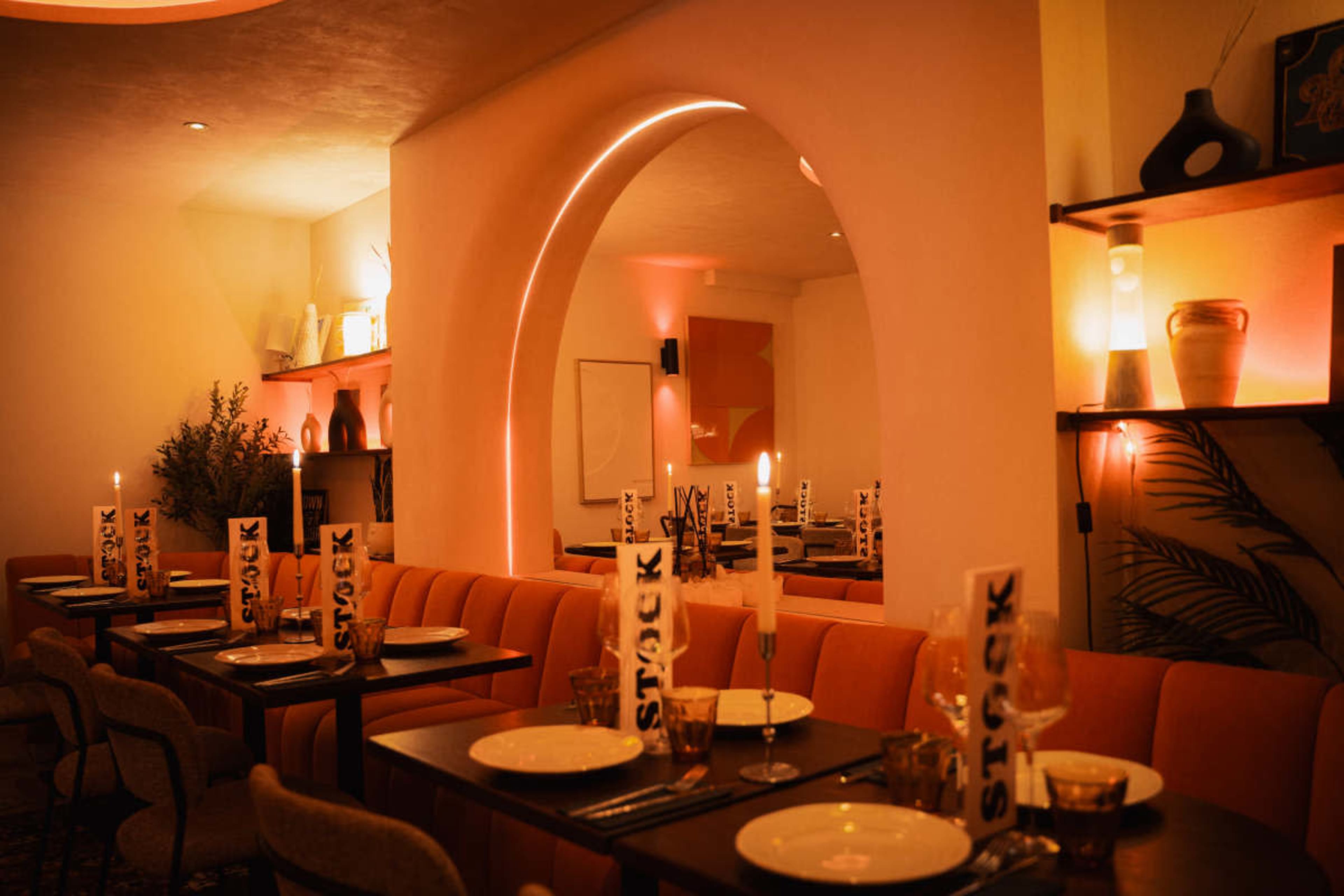 The image shows a cozy restaurant interior featuring orange seating, neatly arranged tables with candles, and warm lighting illuminating the space.