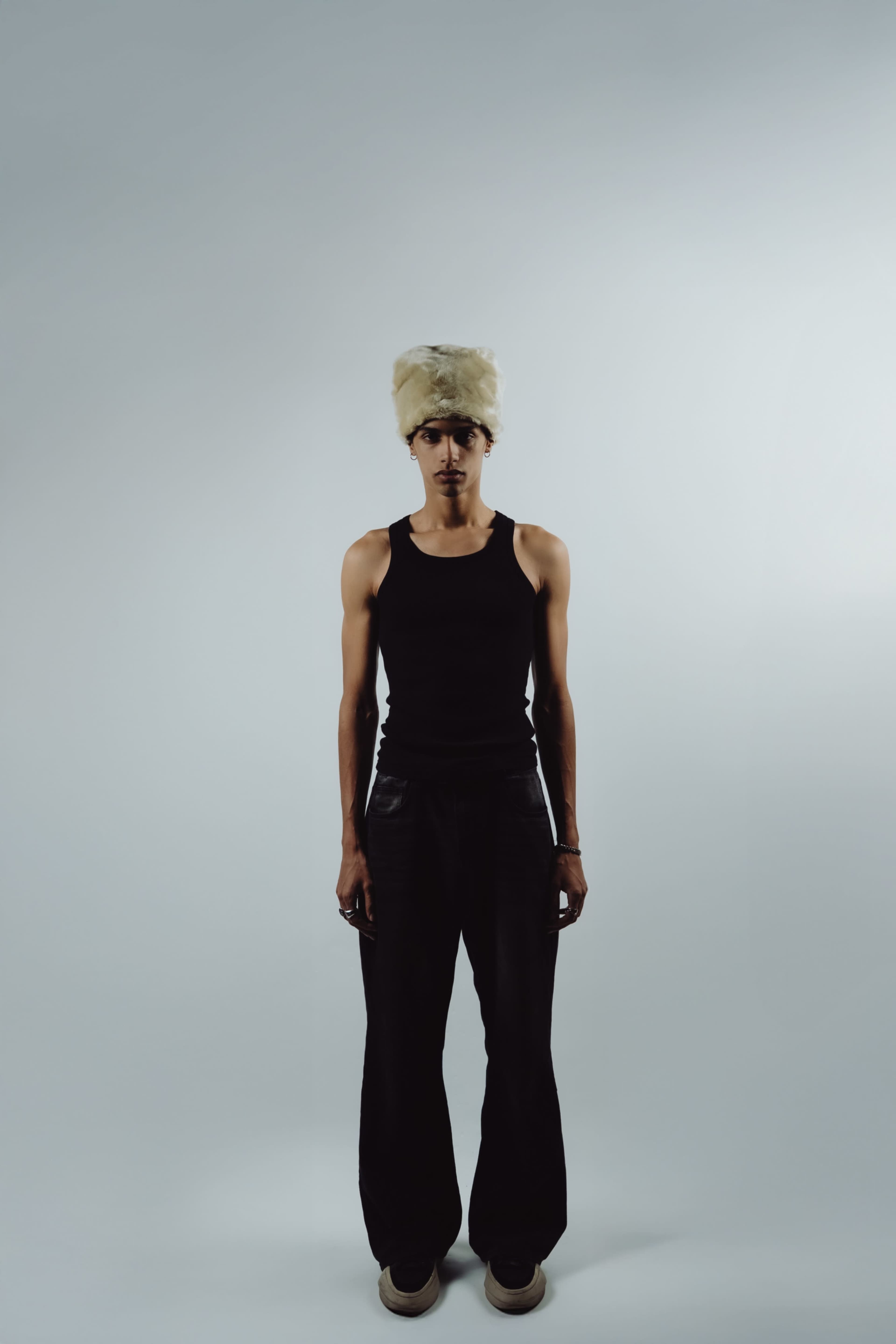 A person stands centrally in front of a plain white backdrop, wearing a black tank top and loose-fitting black pants, with a blonde fur hat.